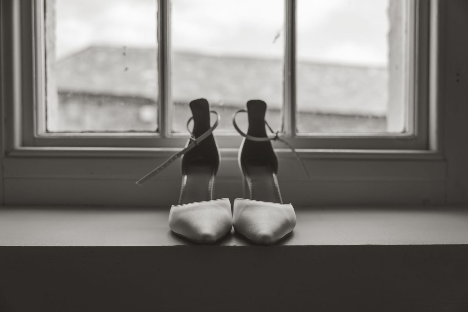 A pair of white wedding shoes sits elegantly on a windowsill with natural light streaming through the window.