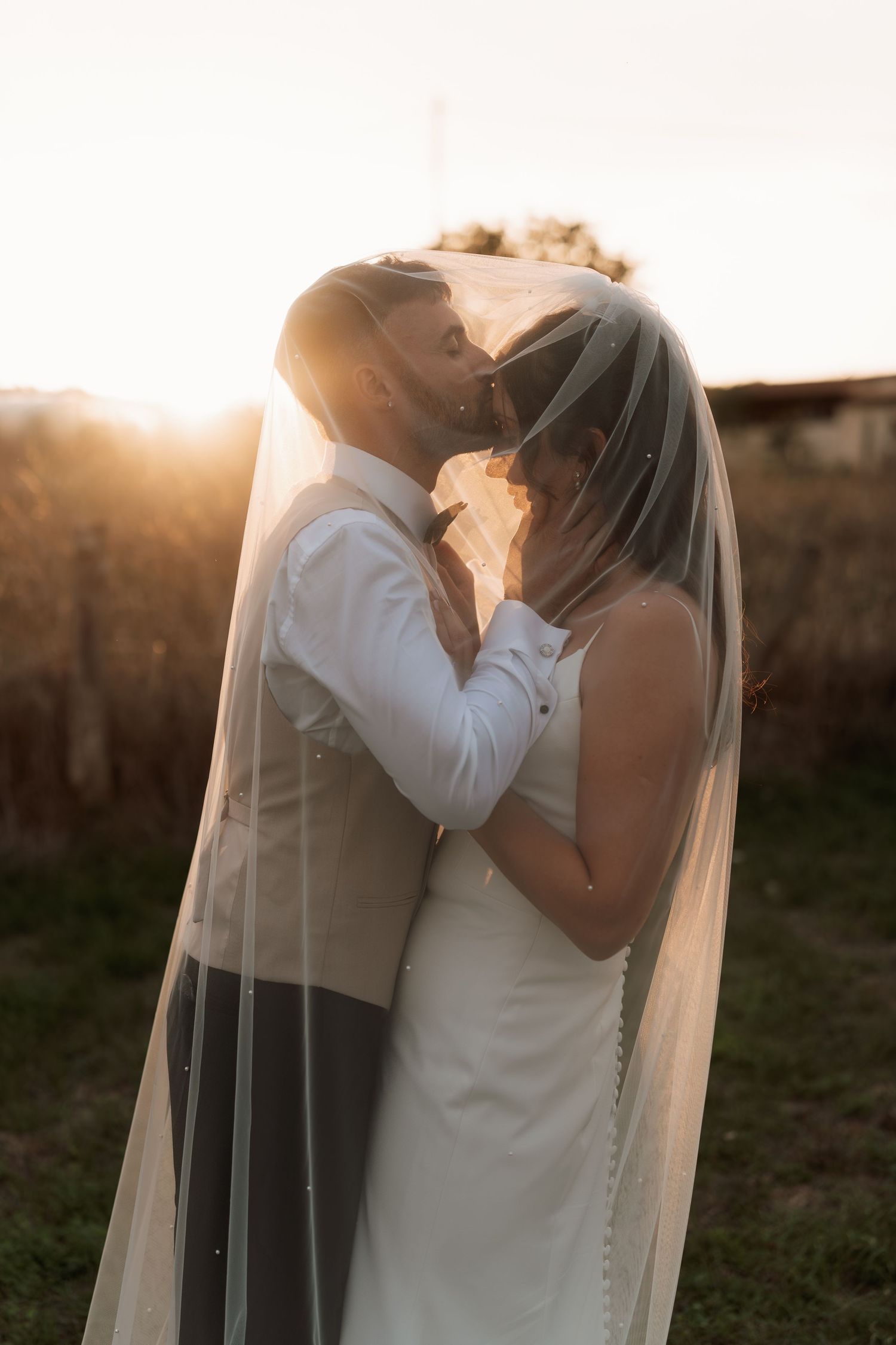 Newlyweds share an intimate moment at sunset with flowing veil in golden light.