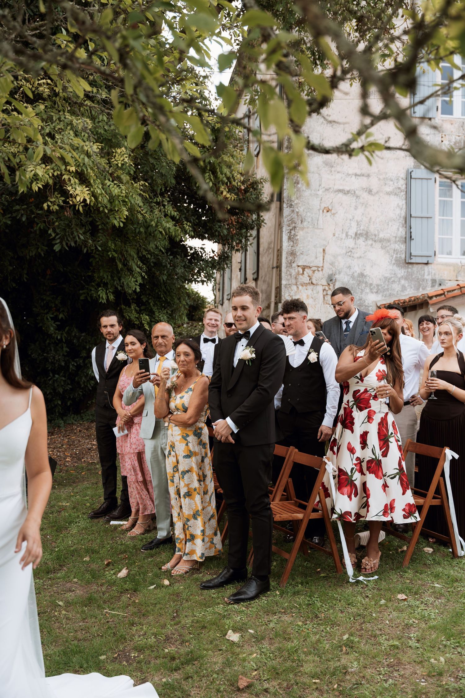 Wedding guests gather outdoors on a grassy lawn next to a stone building for a group photo.