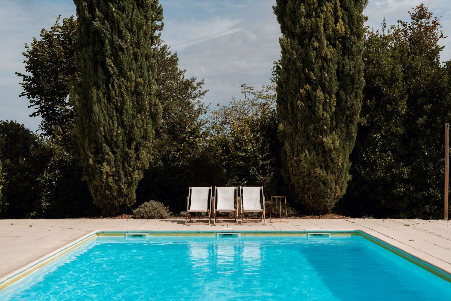 Panoramic view of a bright blue swimming pool surrounded by cypress trees on a sunny day.