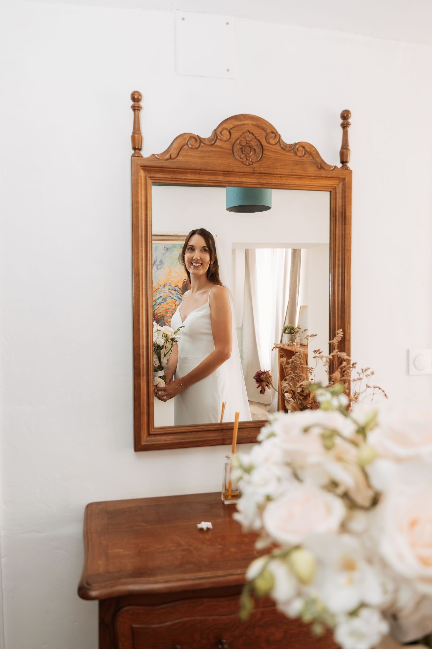 Ornate wooden mirror reflects bridal preparations with white flowers in foreground.