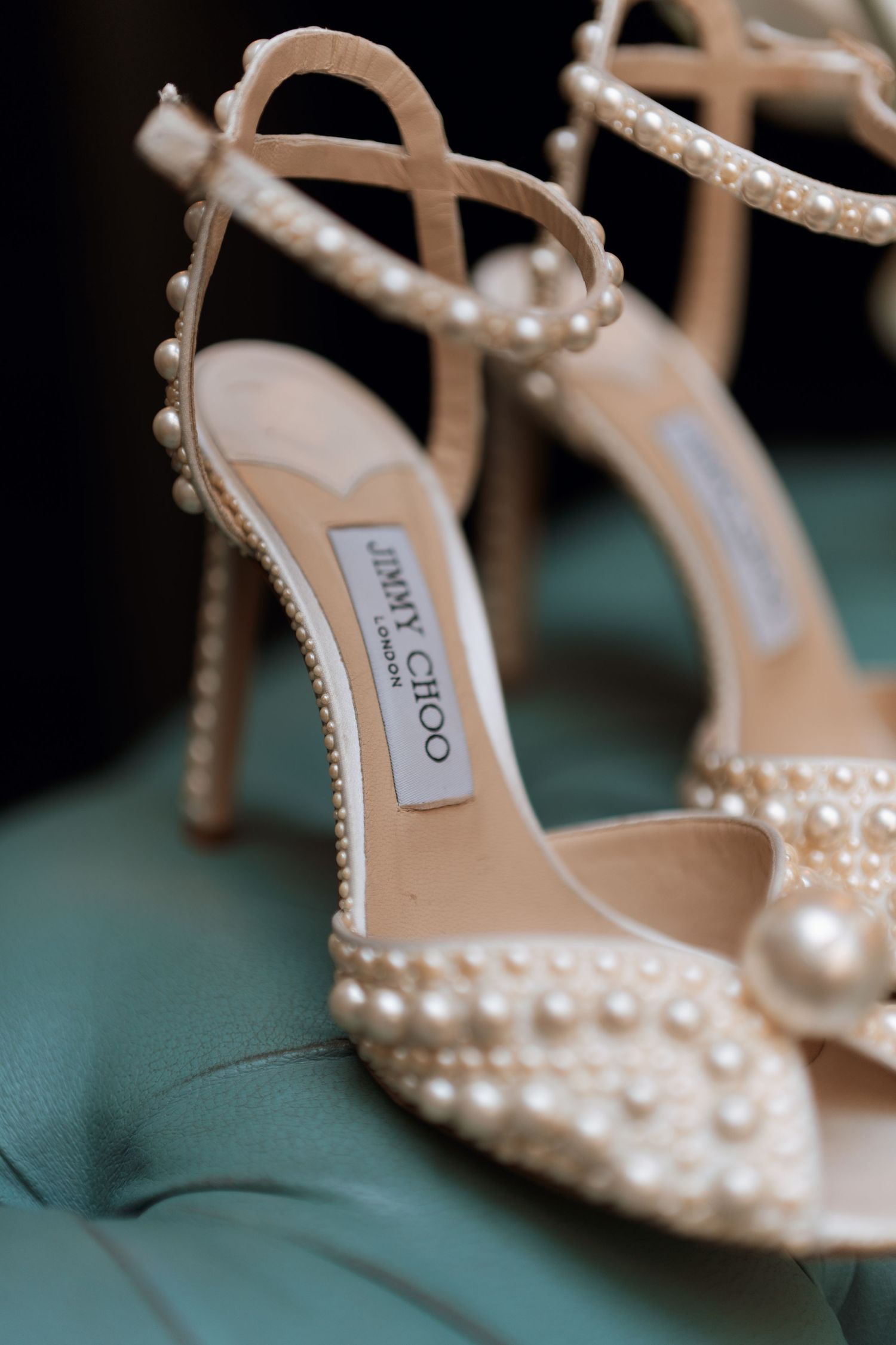 Pearl-studded Jimmy Choo bridal heels with delicate straps displayed against a teal background.