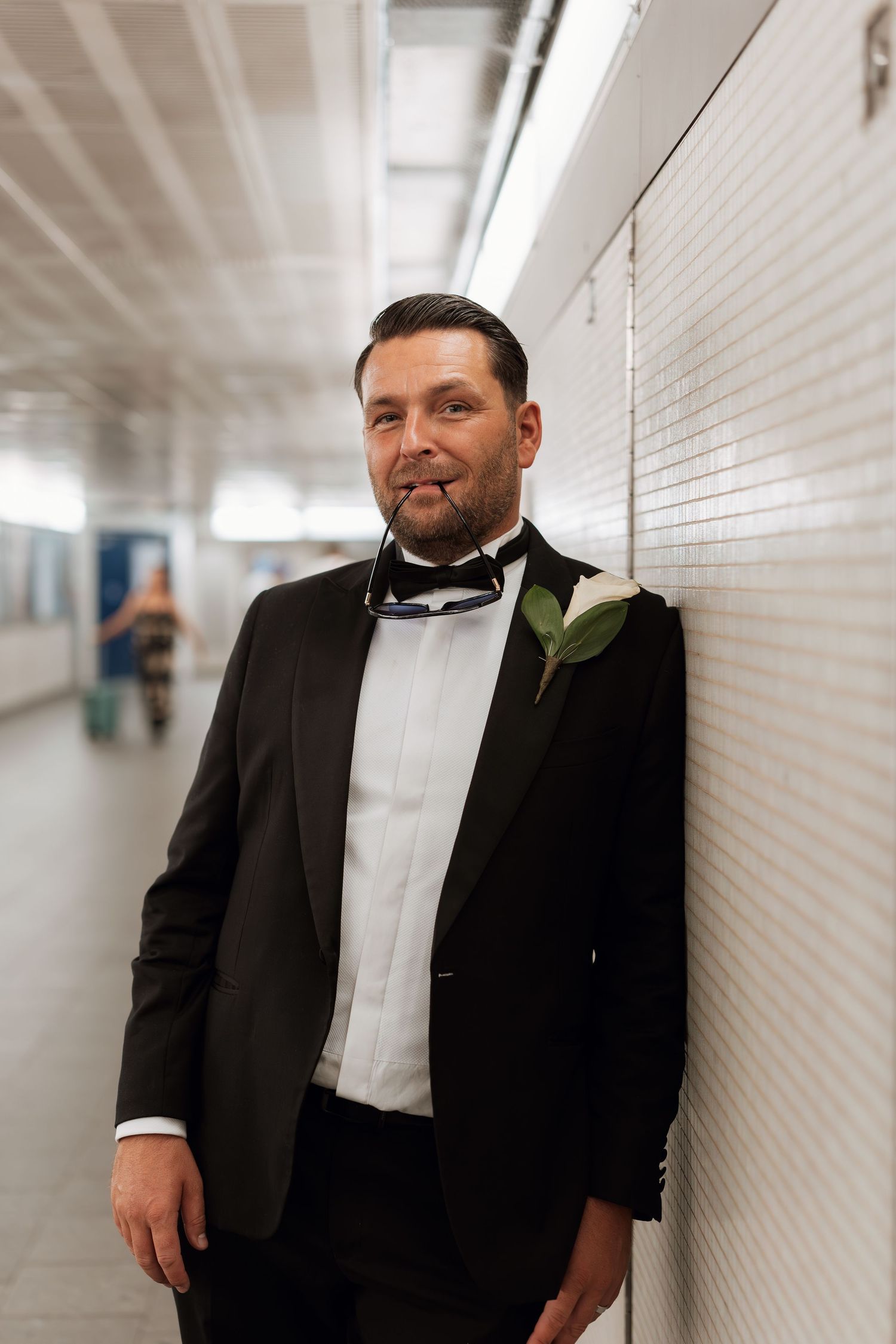 Man in formal black tuxedo and bowtie leaning against white tiled wall in hallway.