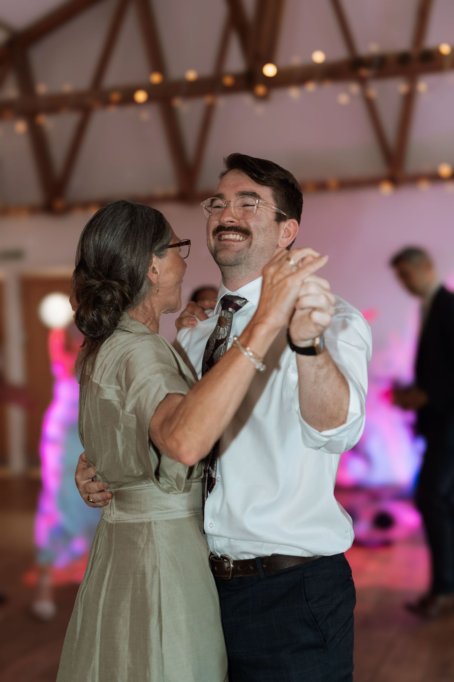 Dancers share a joyful moment during lively dance at reception with pink lighting in background.