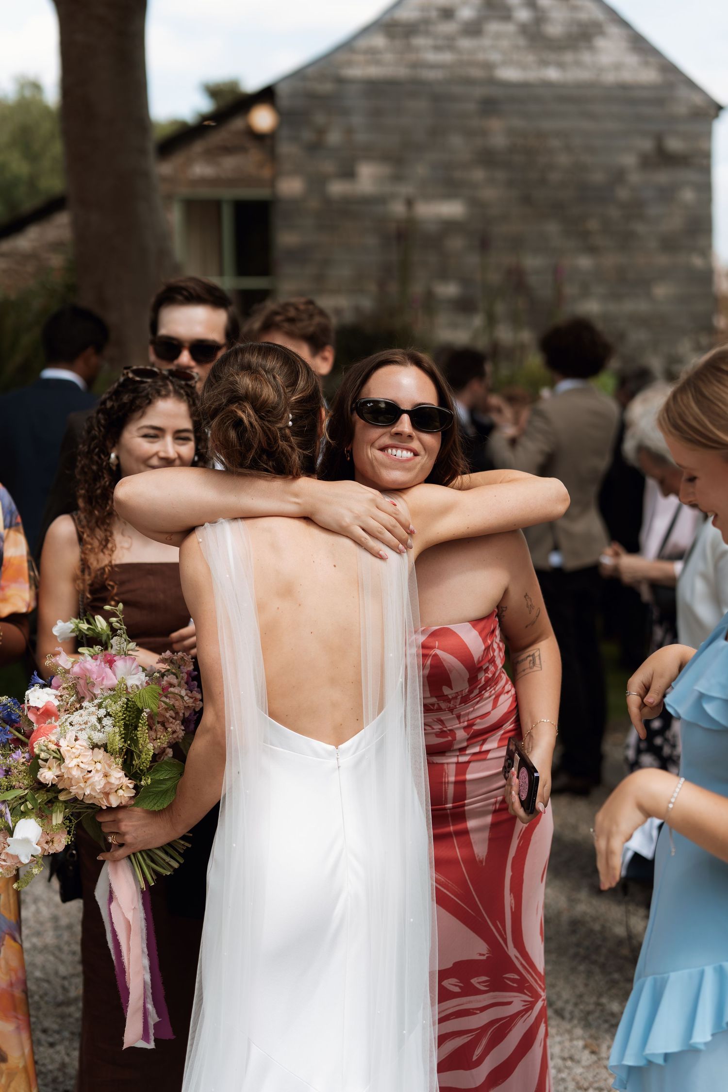 Wedding guests gathered outside a rustic building sharing embraces and congratulations.