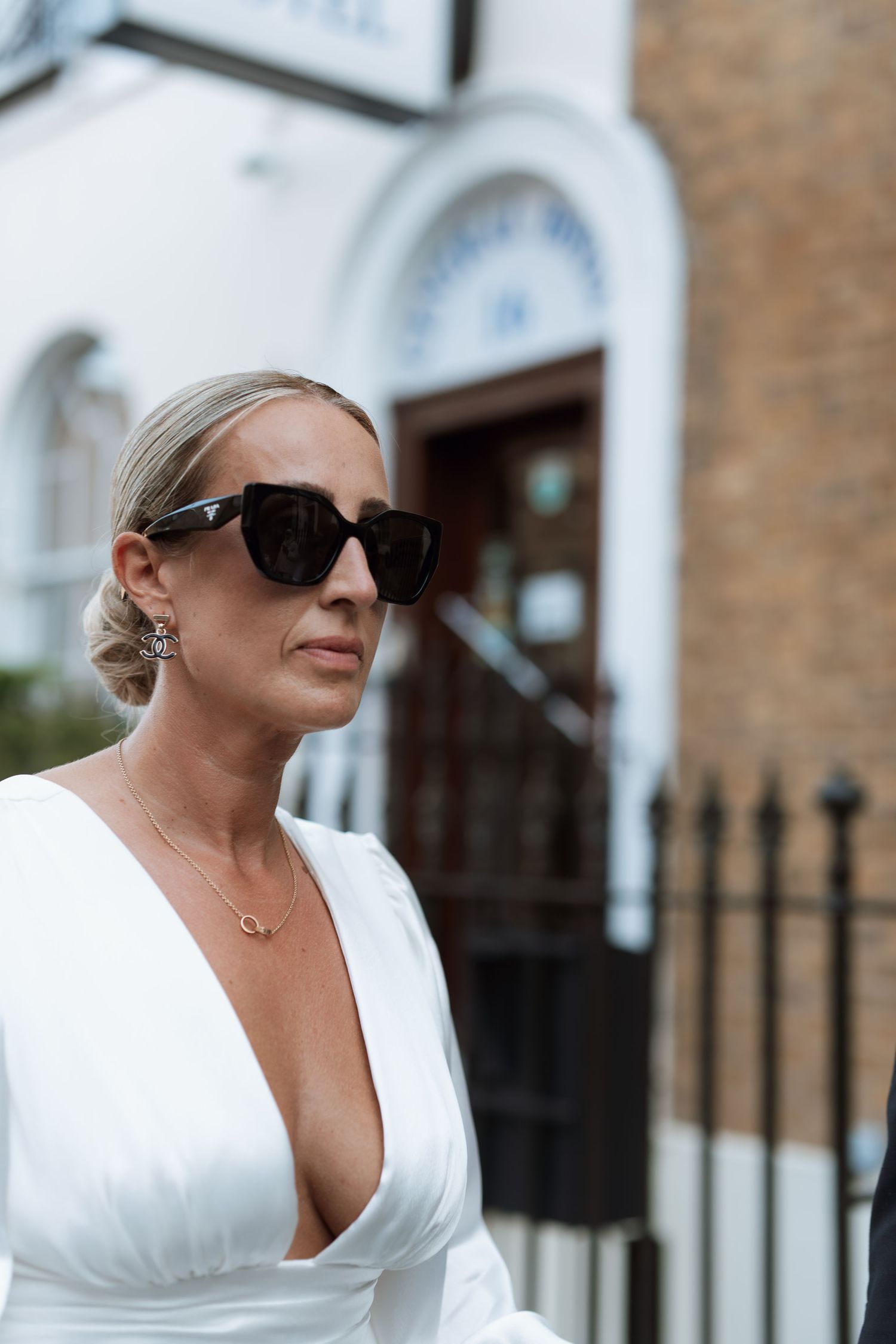 Woman in white dress and sunglasses walking along a brick building exterior.