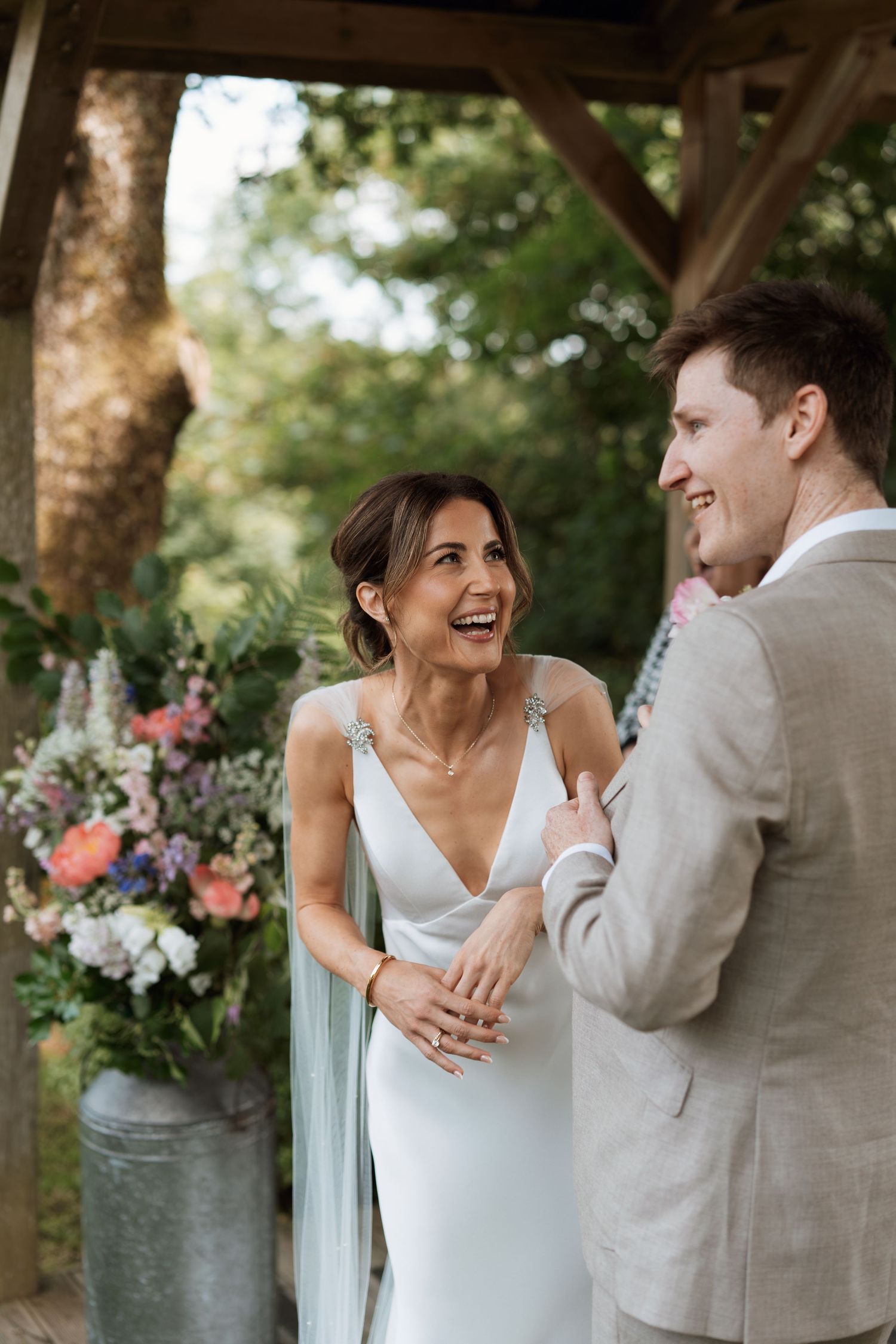 Joyful wedding couple sharing a laugh under wooden pergola with floral decorations.