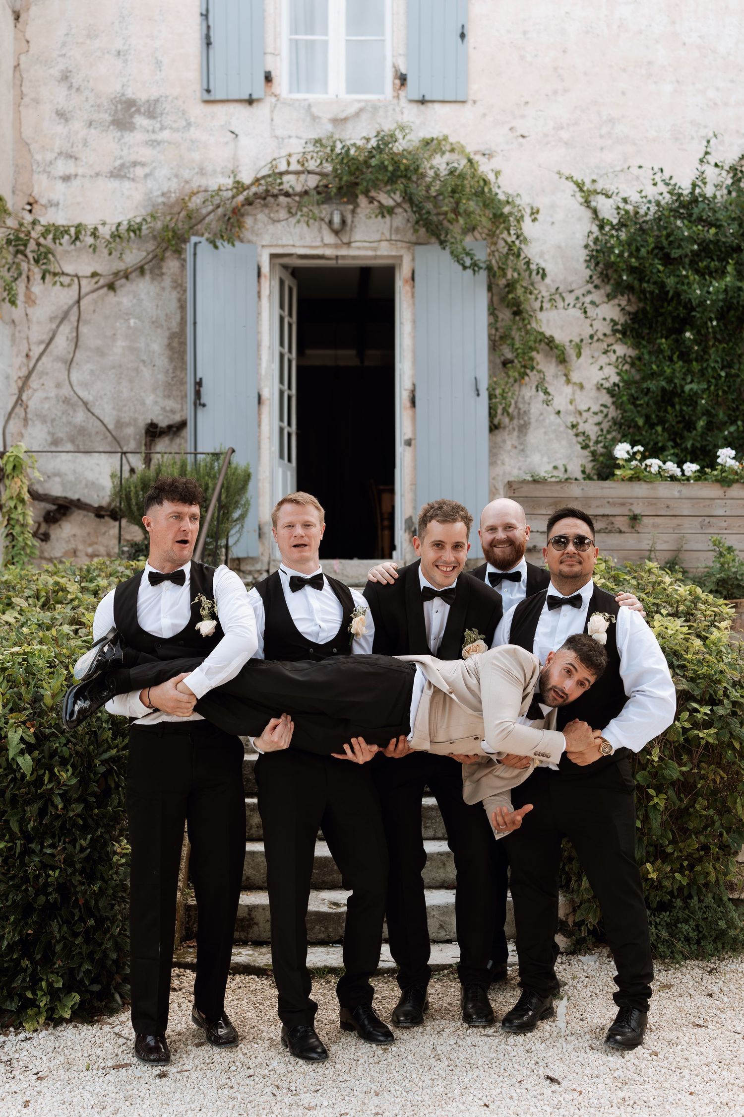 Groomsmen in black and white attire sit casually on a stone wall for a group photo.