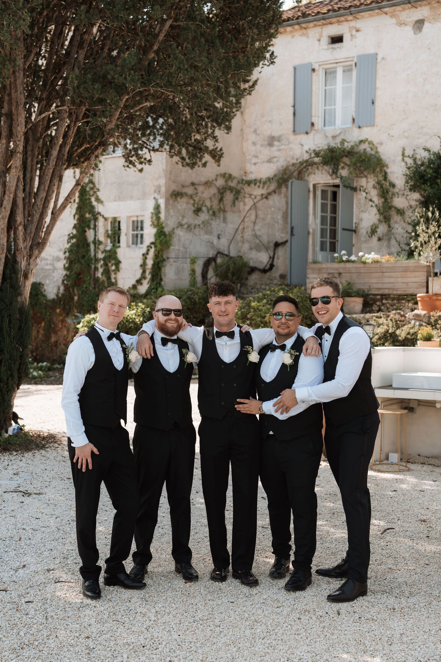 Group photo of groomsmen in matching black vests and bow ties standing outside a white building.