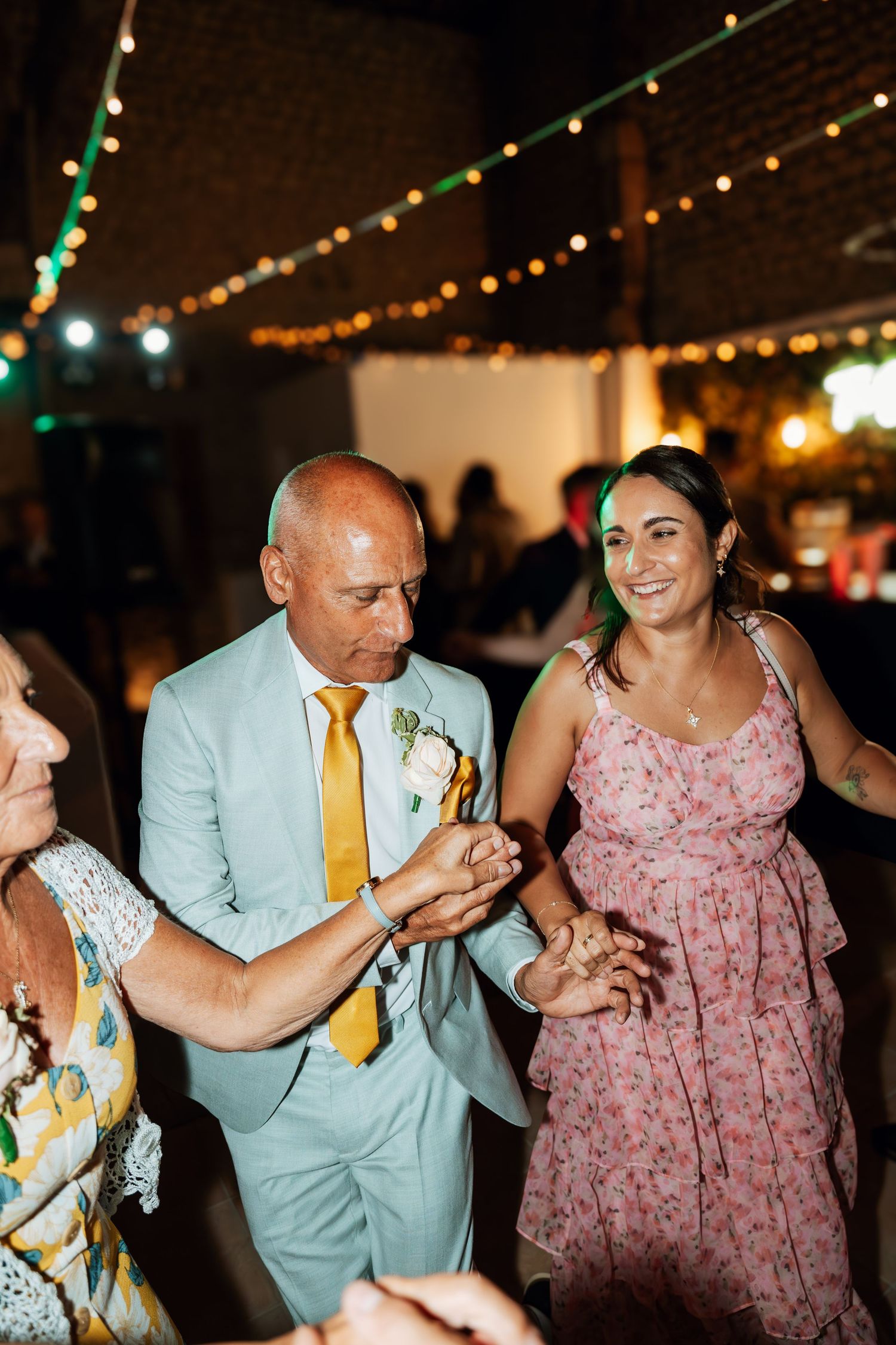 Wedding guests dancing in formal attire under string lights at an evening reception.
