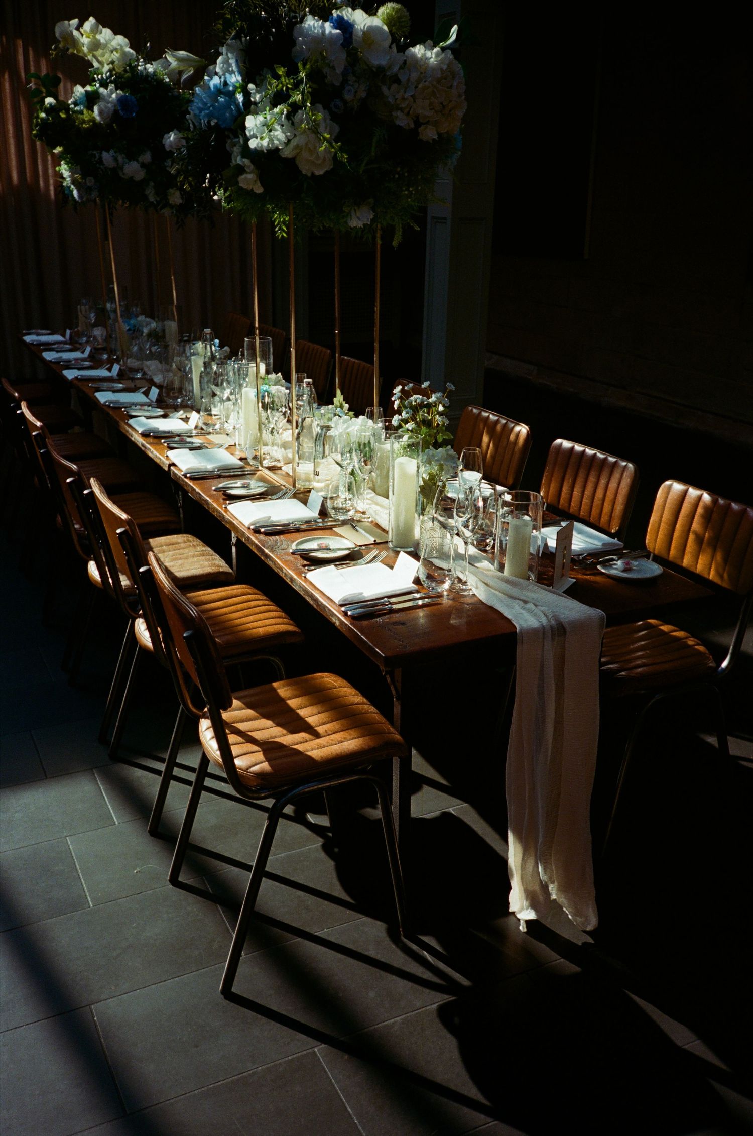 Elegant wedding table setting with wooden chairs, white floral arrangements and flowing fabric in dramatic lighting.