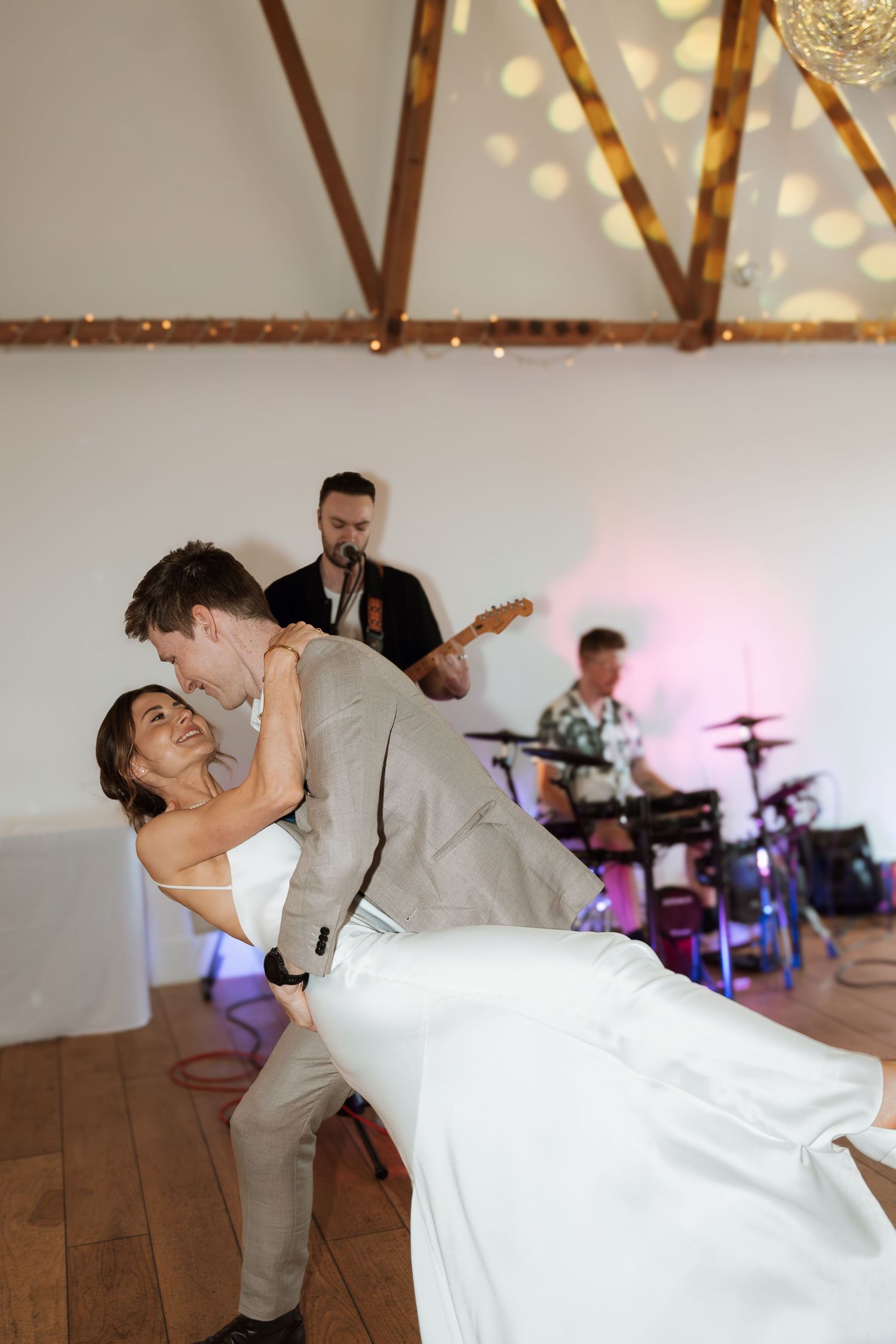 Couple performs romantic dance dip on wooden floor with string lights overhead during wedding reception.