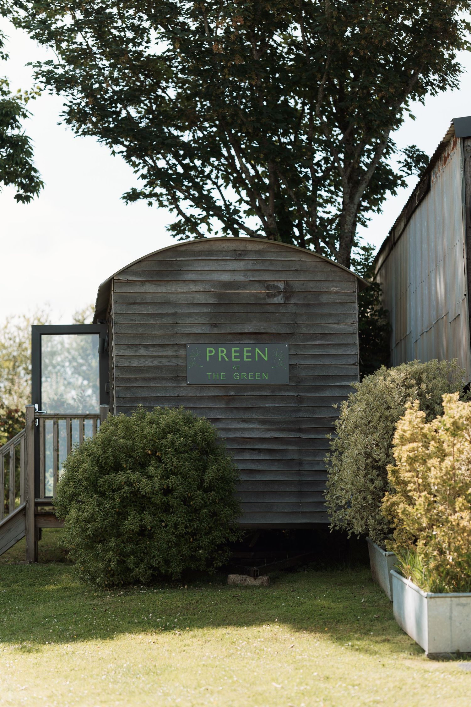 A rustic wooden shed with a green 'PREEN' sign surrounded by lush greenery and bushes.