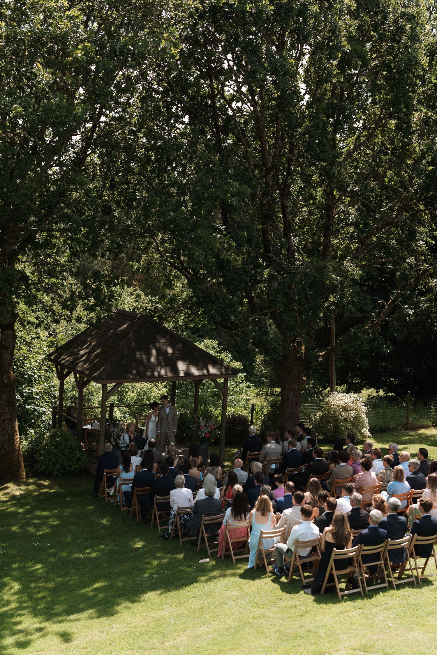Aerial view of wedding guests seated at an outdoor ceremony under a wooden gazebo surrounded by trees.