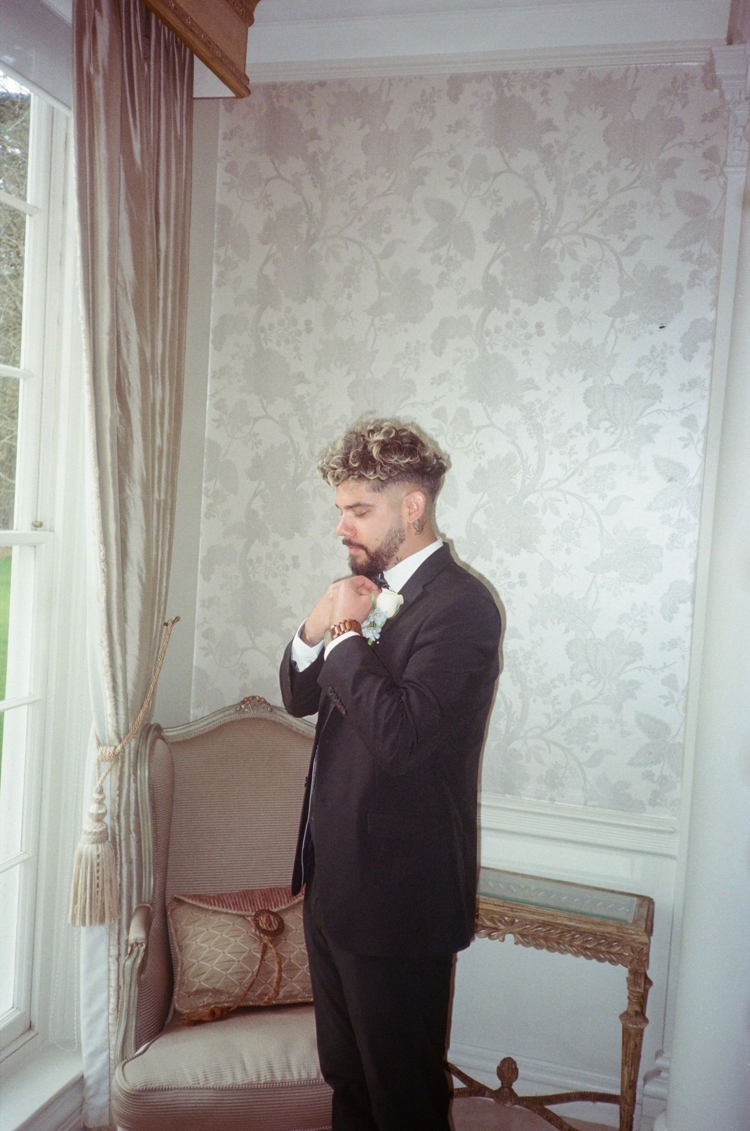 A person in formal attire stands in contemplation by a window in an elegant room with light curtains and textured walls.