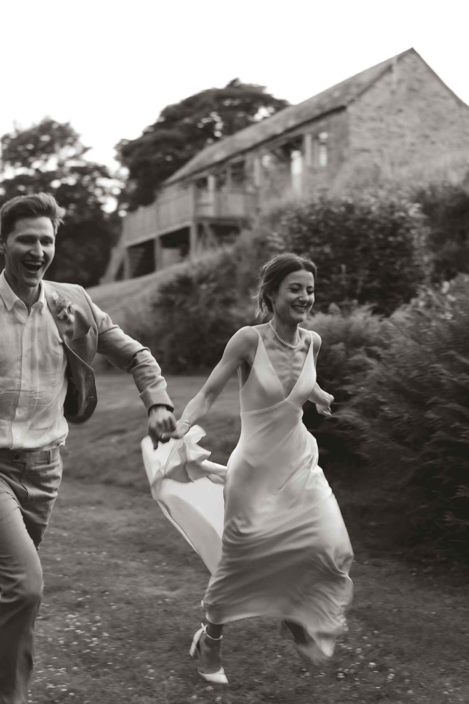 Black and white photo of couple running joyfully outside stone building during wedding celebration.