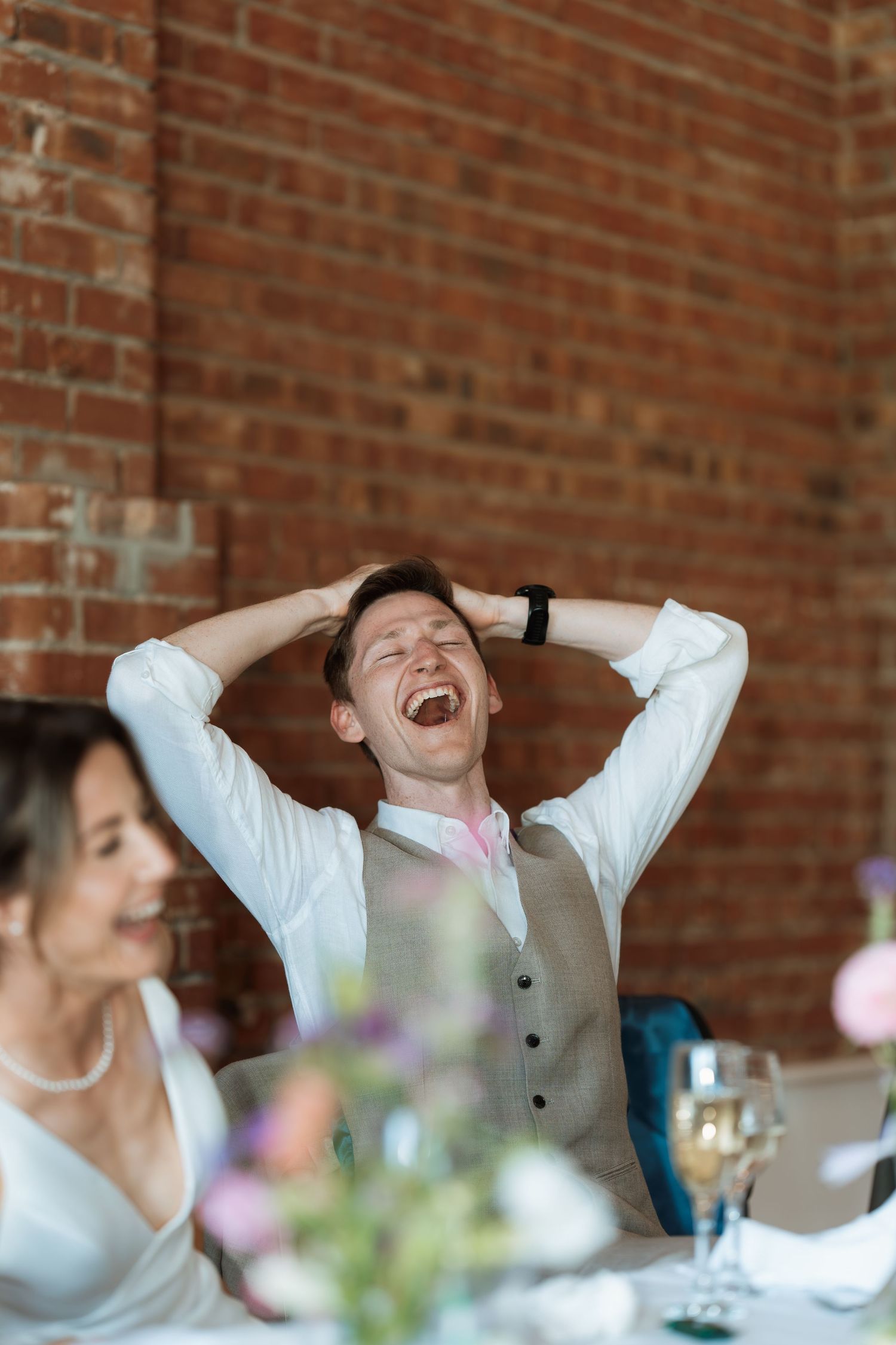 A joyful guest at a wedding reception laughs heartily while seated at a decorated table.
