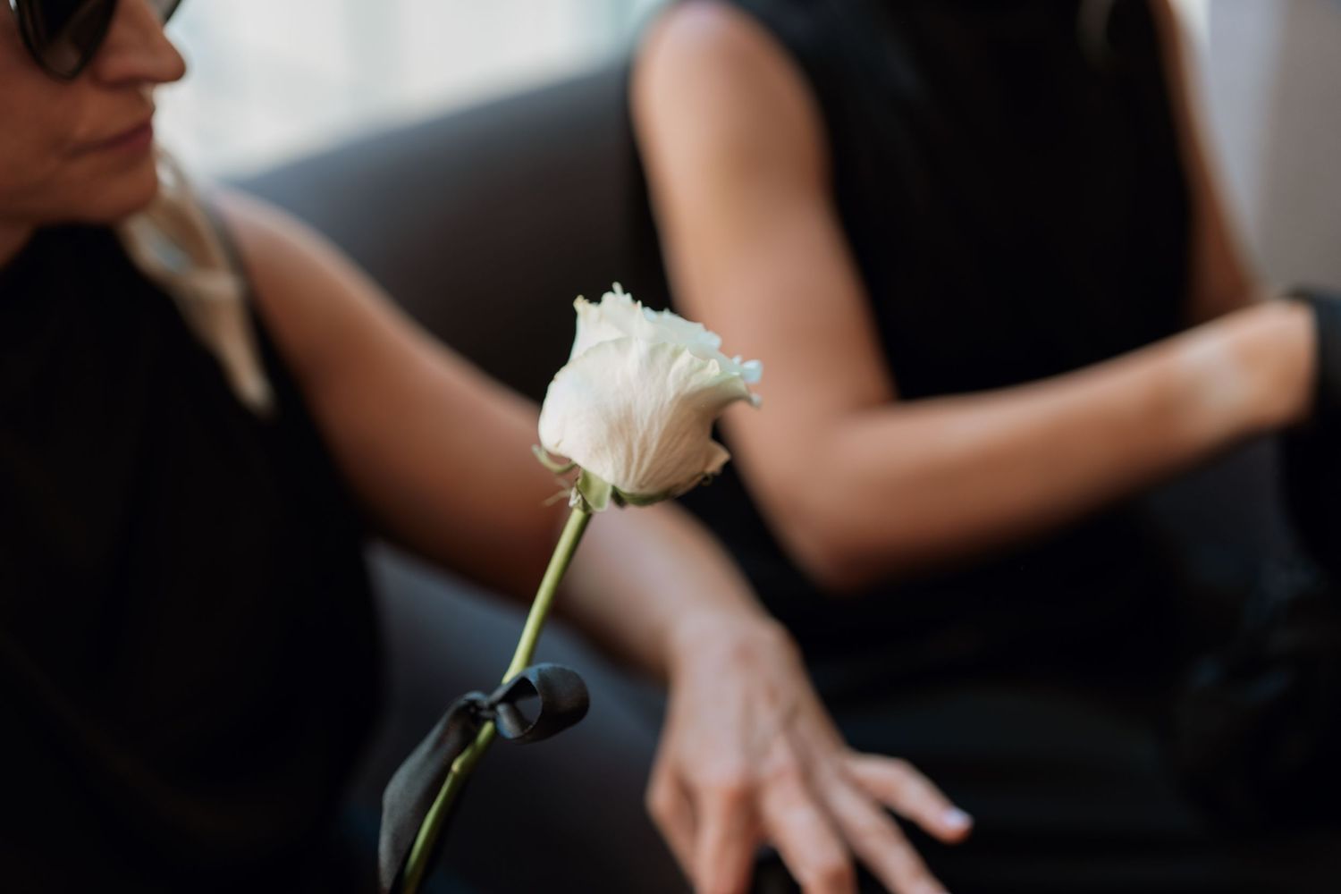 A person wearing a black outfit holds a white flower while standing in a dark setting.
