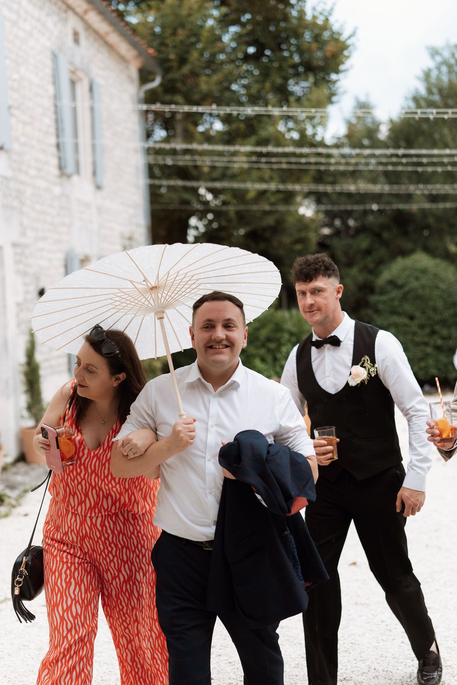 Wedding guests walk together outdoors, with one holding a decorative white parasol.