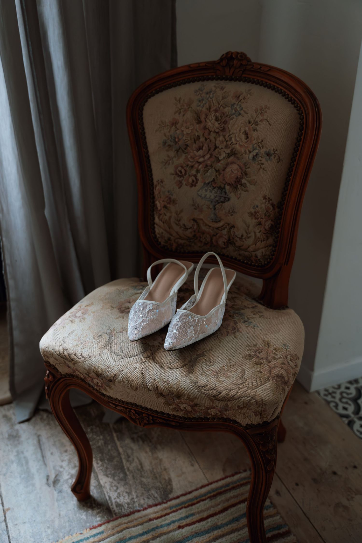 Elegant white lace bridal shoes displayed on vintage floral upholstered chair with ornate wooden frame.