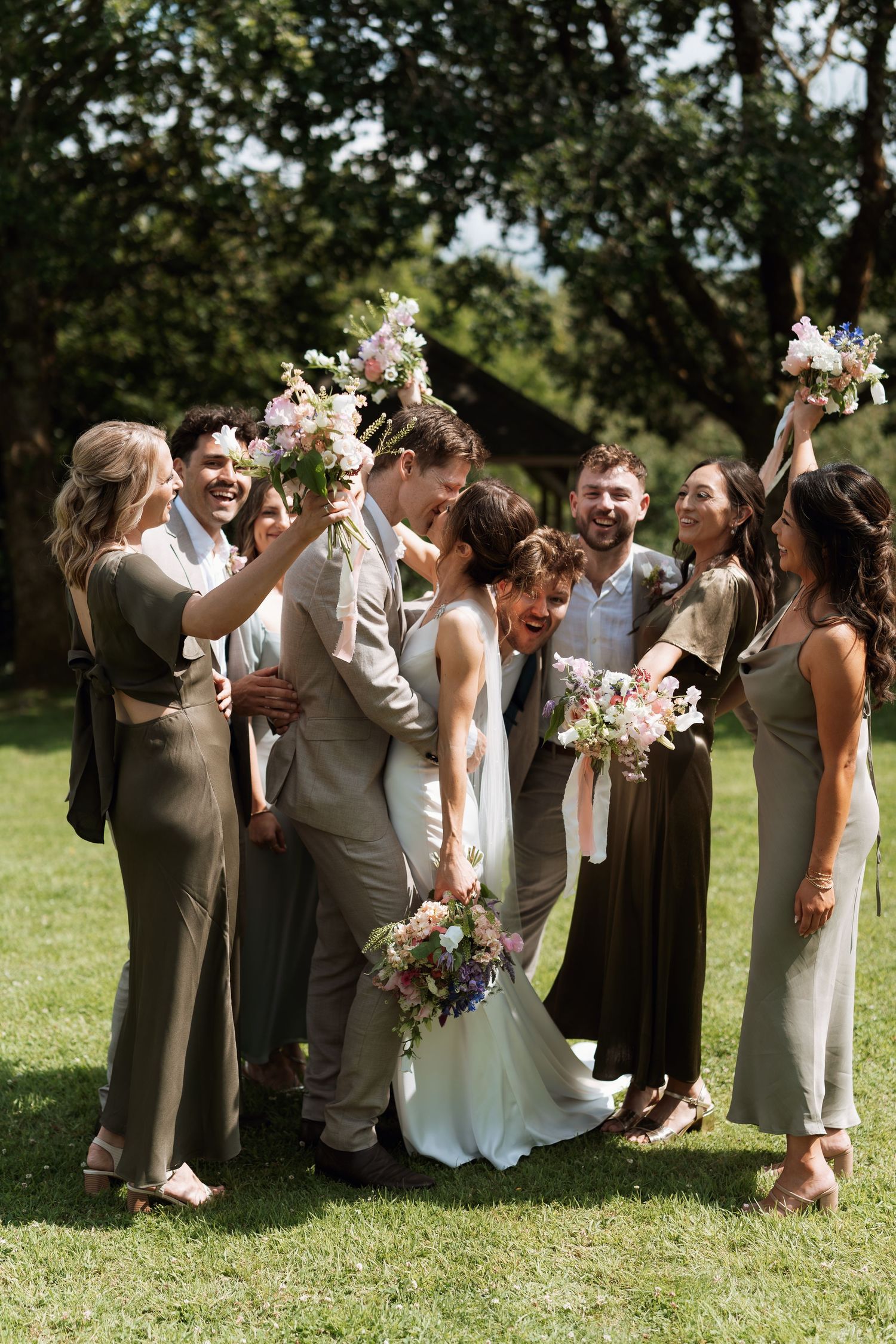 Wedding party poses playfully on a grassy lawn with bridesmaids in dark dresses and groomsmen in light vests.