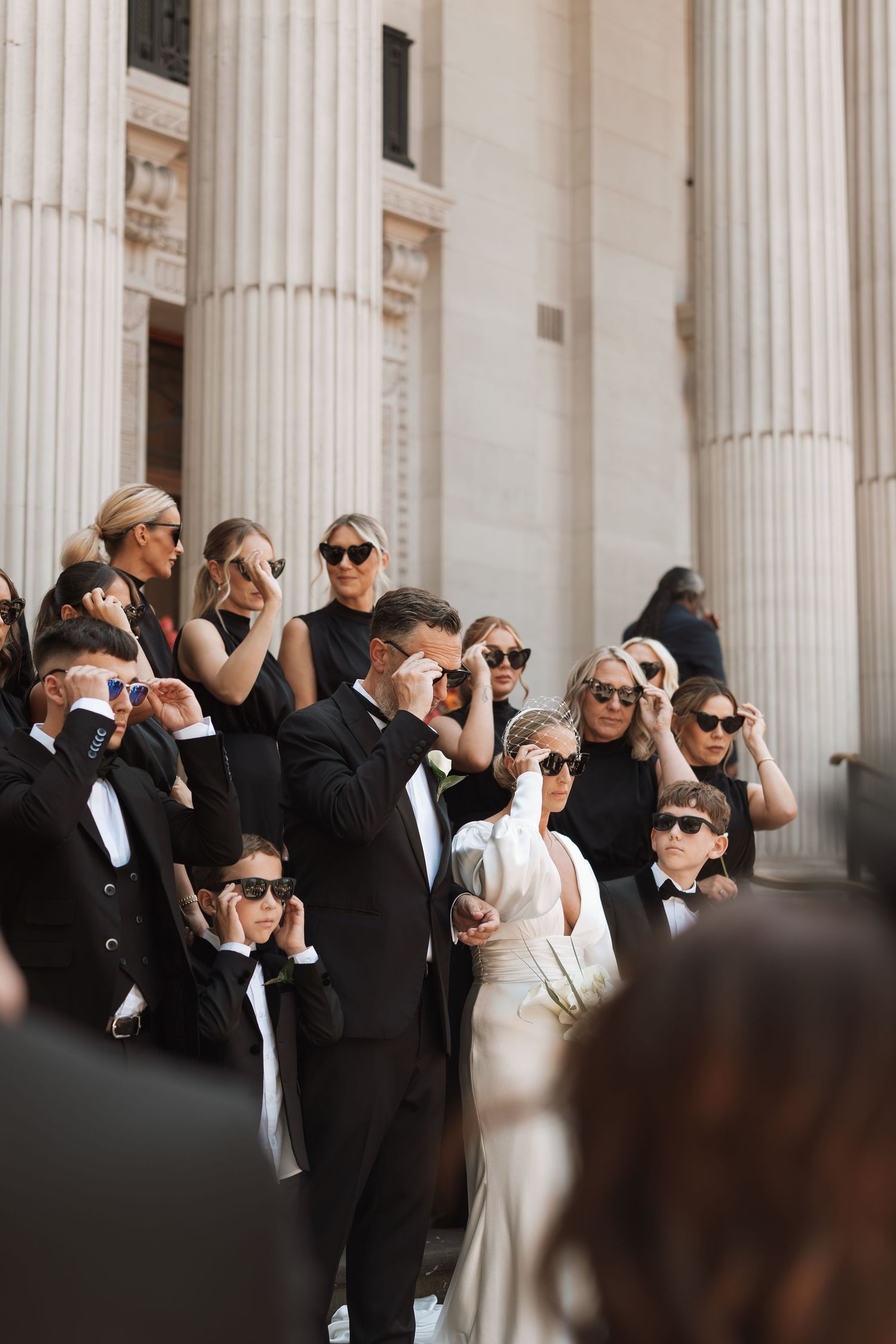 Wedding guests in formal wear gather outside a columned building taking photos.