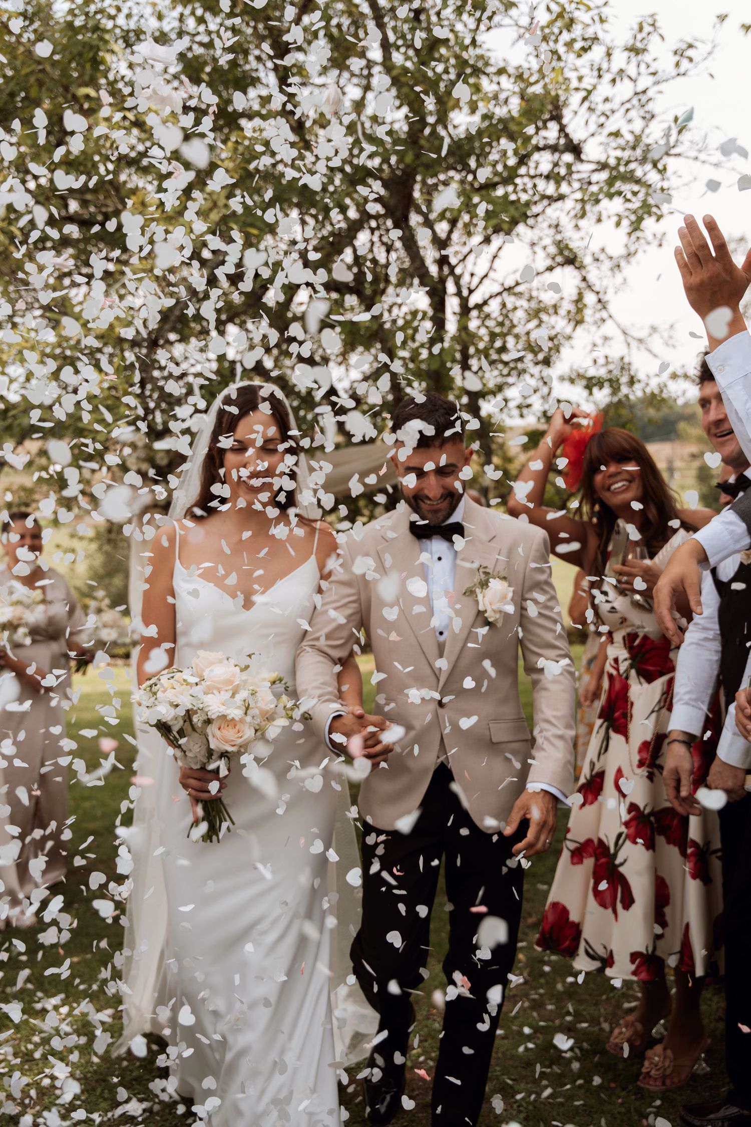 Wedding couple and guests celebrate with confetti toss under trees during outdoor ceremony.