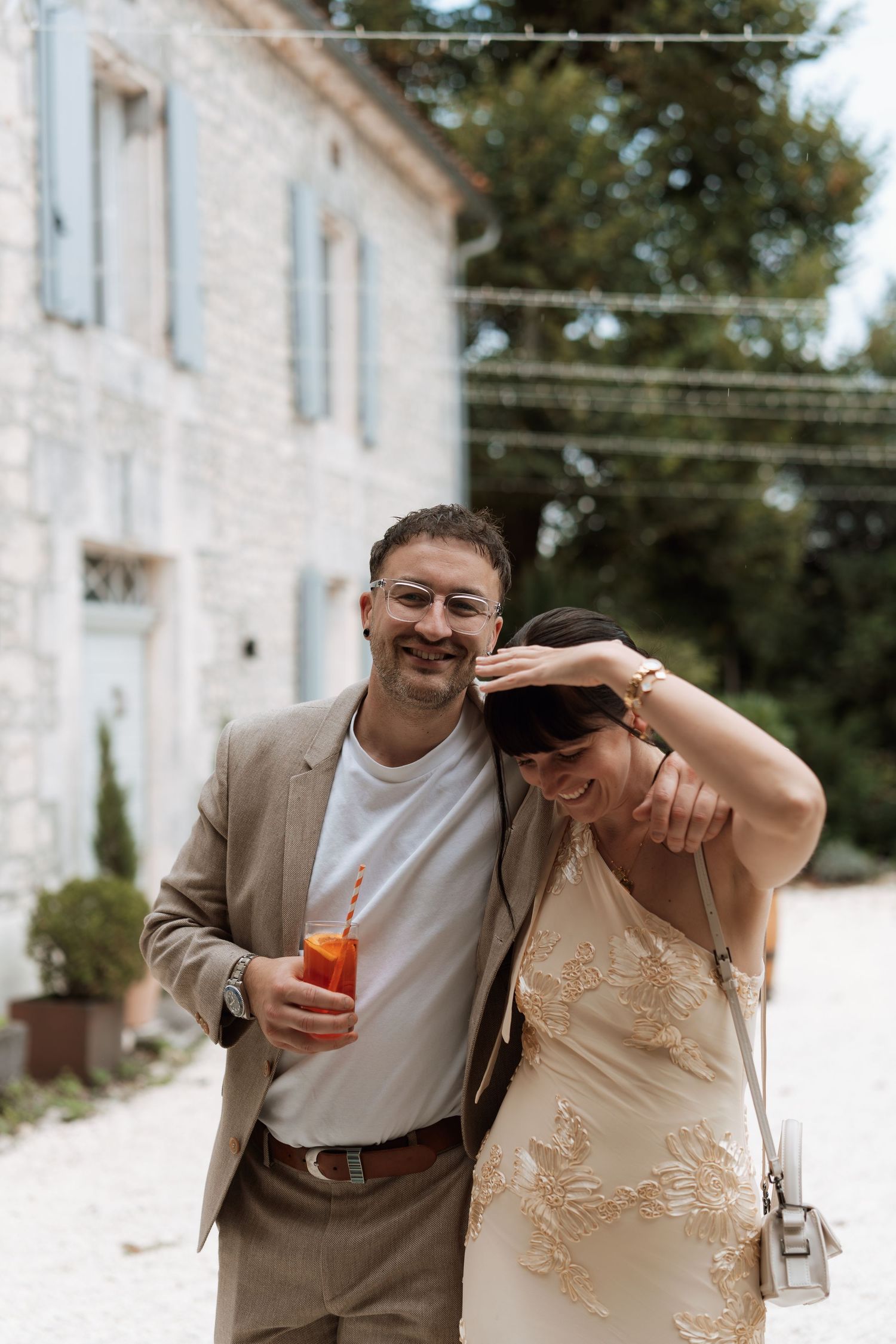 A couple shares a candid moment while enjoying drinks outside a white stone building.