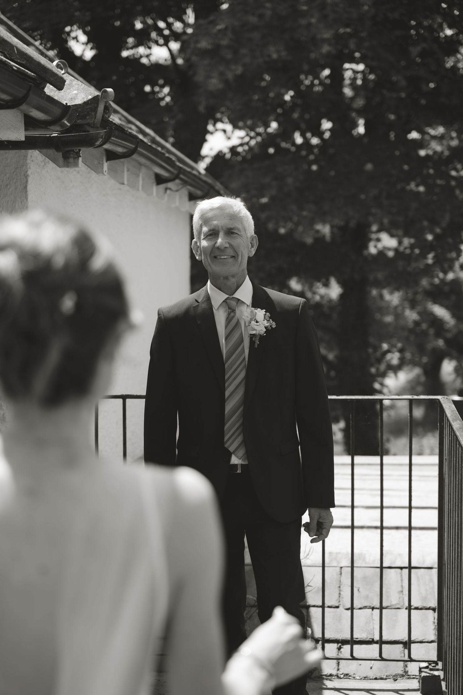 Black and white photo of a groom in a suit and tie standing at an outdoor wedding venue.