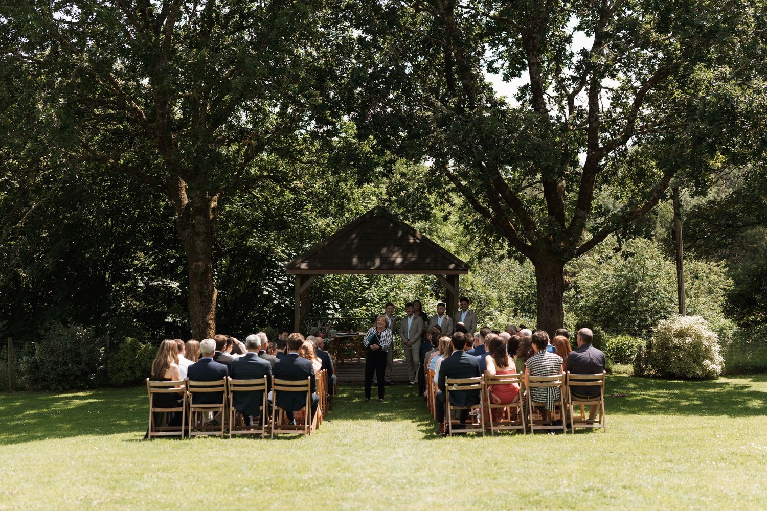 Outdoor wedding ceremony setup with wooden chairs arranged under large trees in a garden setting.