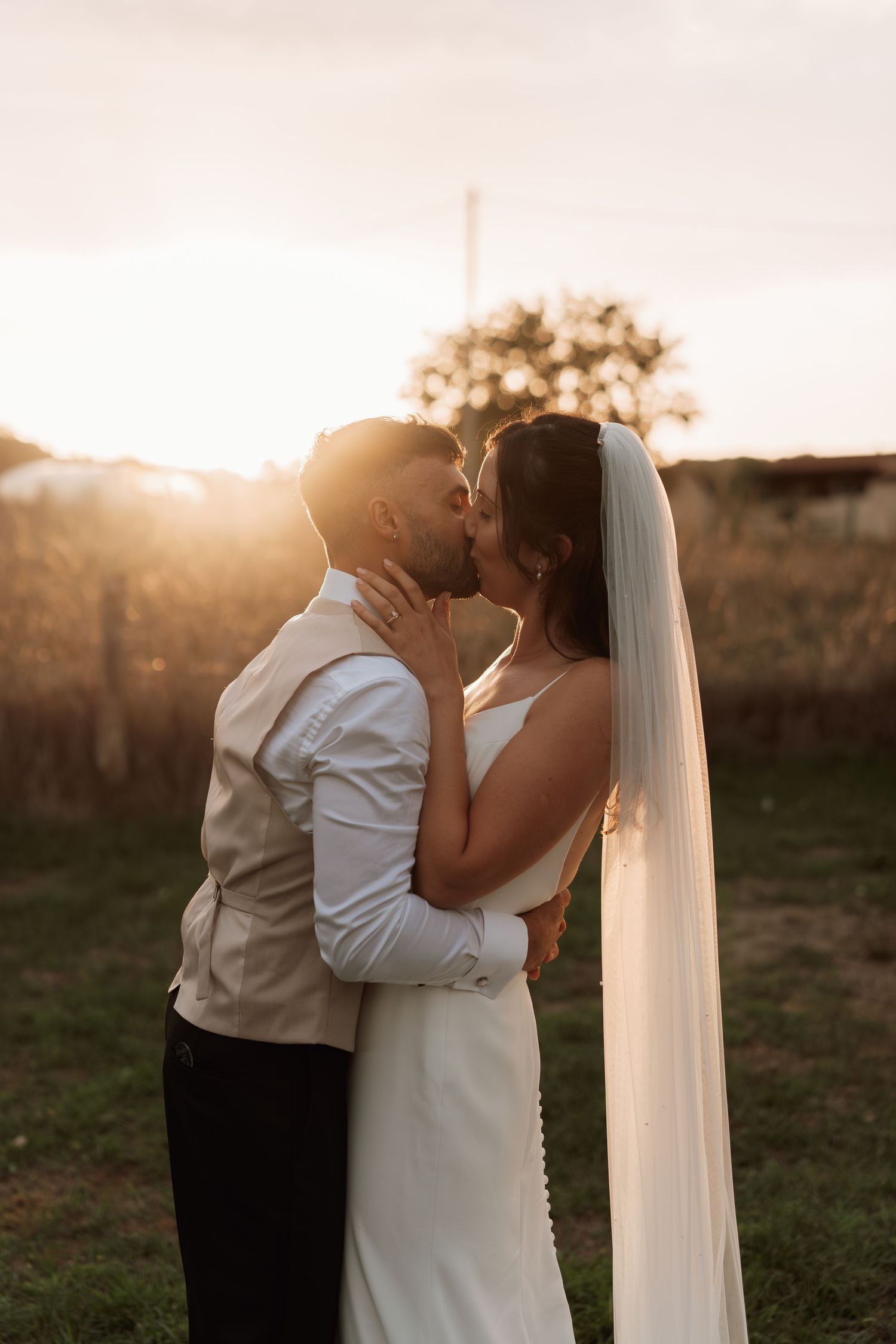 Silhouetted couple sharing intimate moment at sunset with bride's veil flowing in golden light.