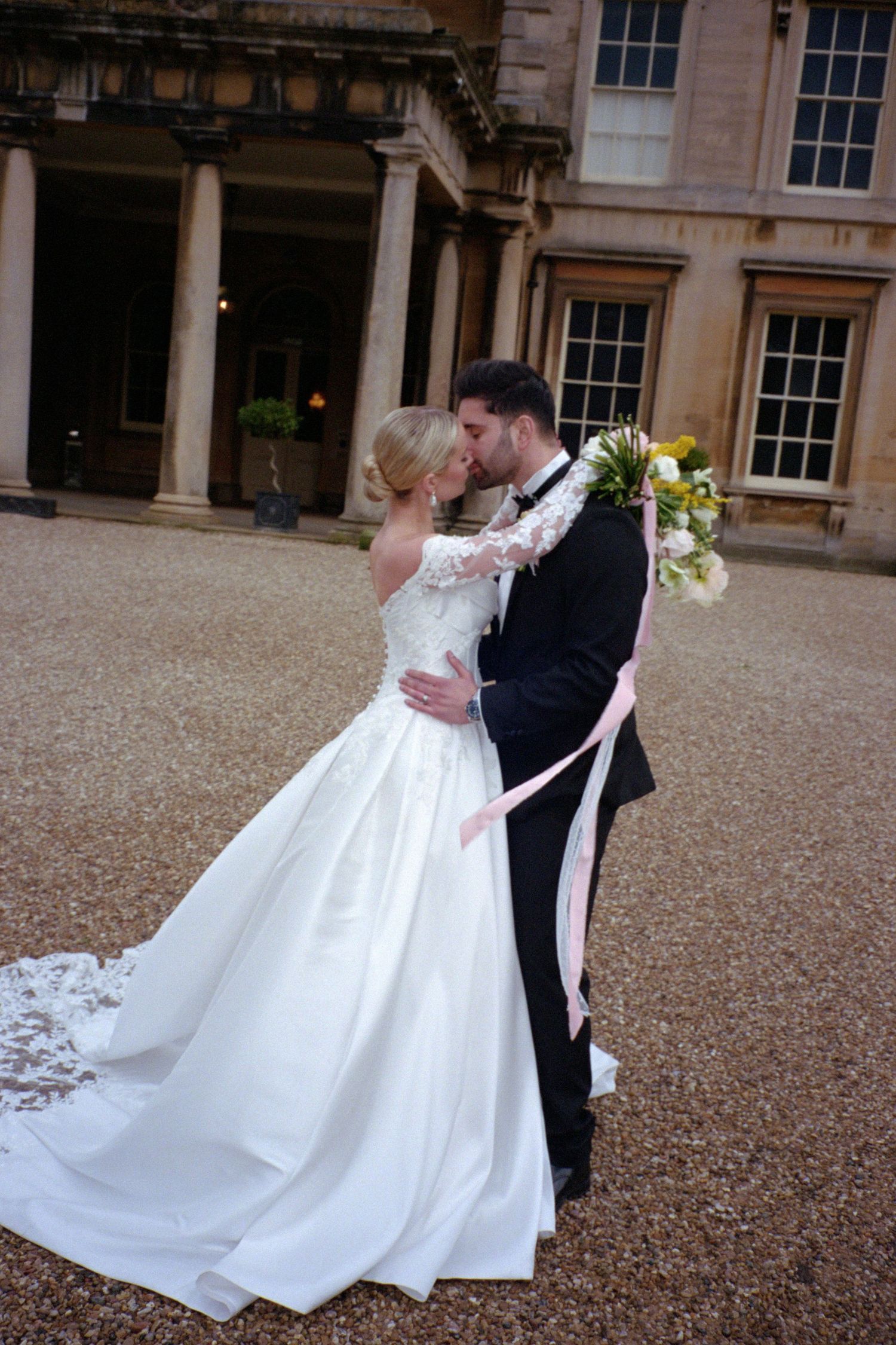 A romantic wedding moment captured outside a grand historic building with classical columns and gravel courtyard.