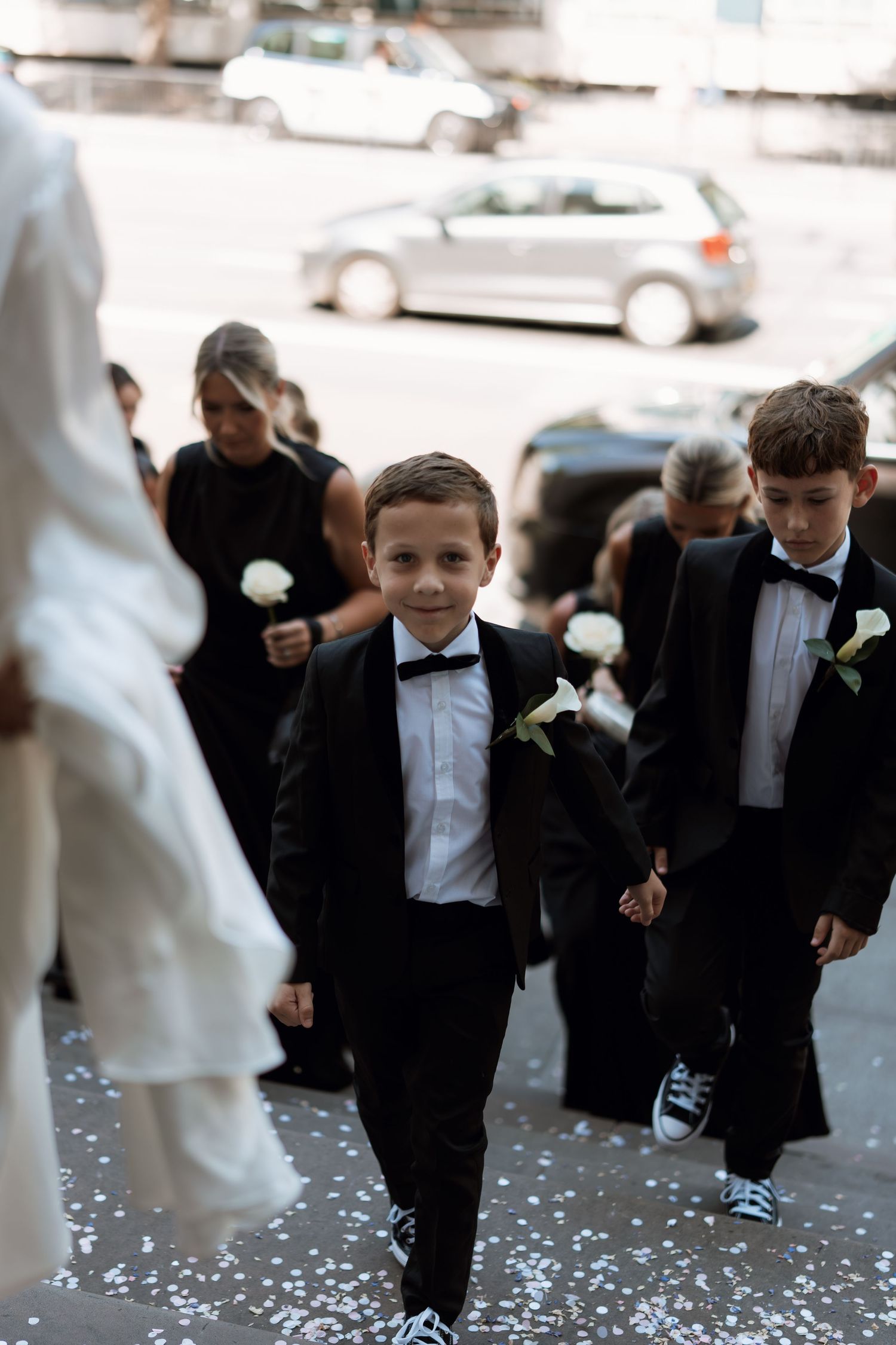 Young ring bearers in black tuxedos walking on a path scattered with white petals.