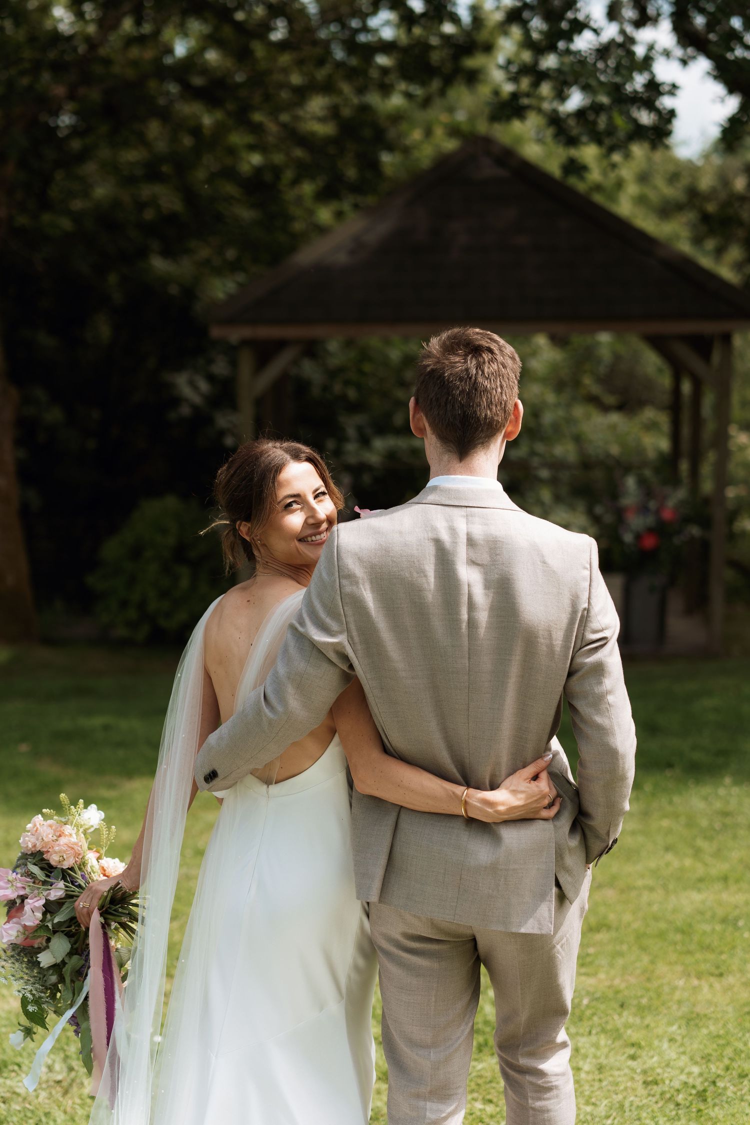 Newlyweds walking together on grass with bride in backless dress holding colorful bouquet.