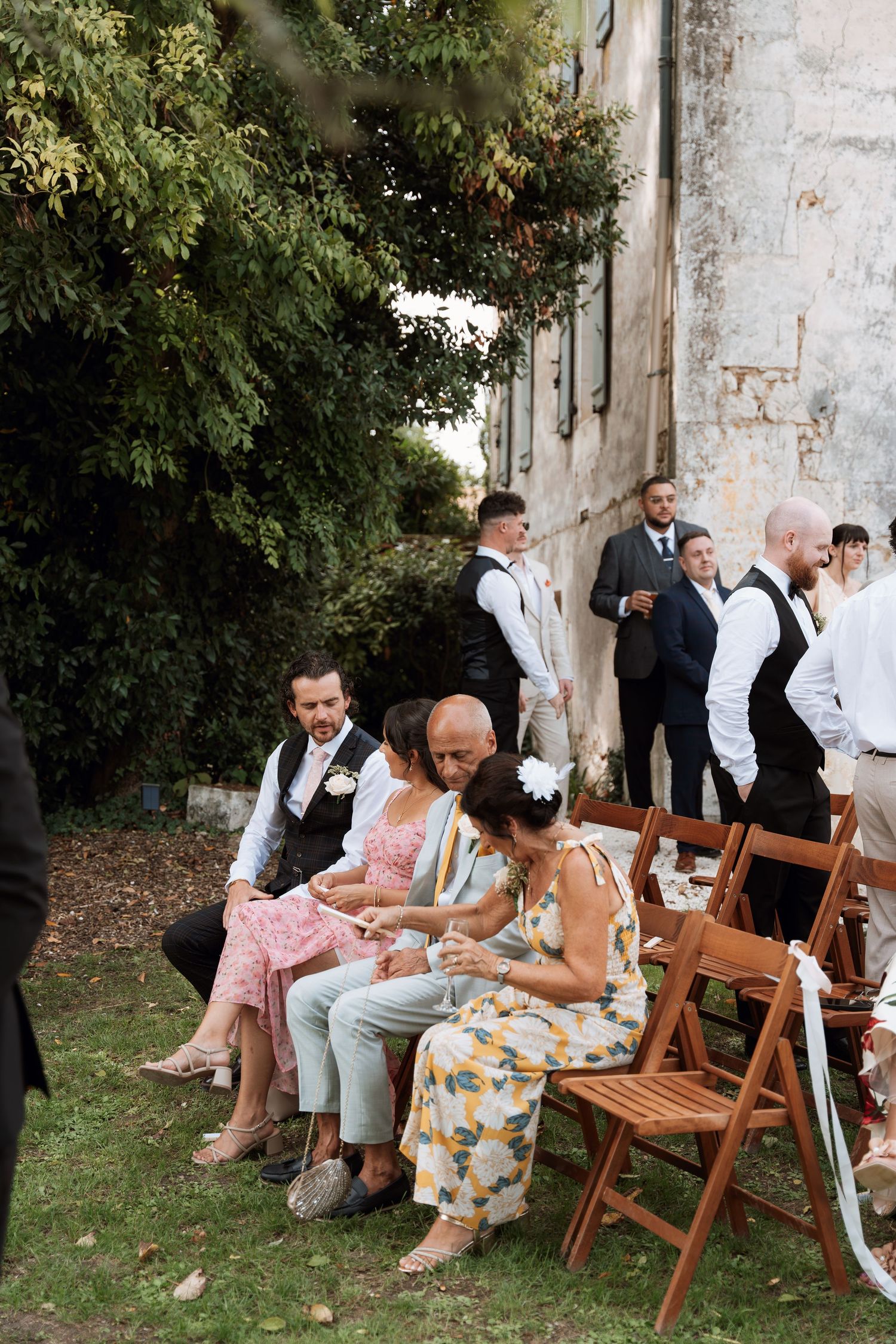 Wedding guests relax on wooden chairs in an outdoor garden setting beside a rustic stone wall.