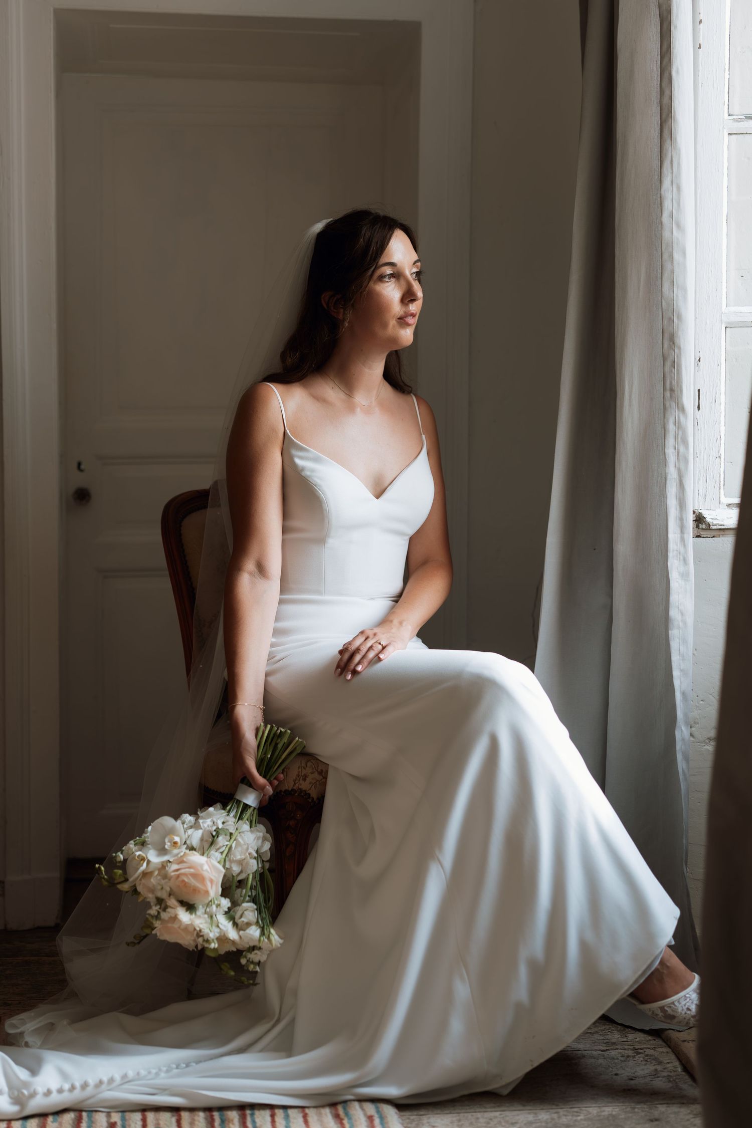 A white wedding dress flows elegantly as someone poses by a window in natural light.