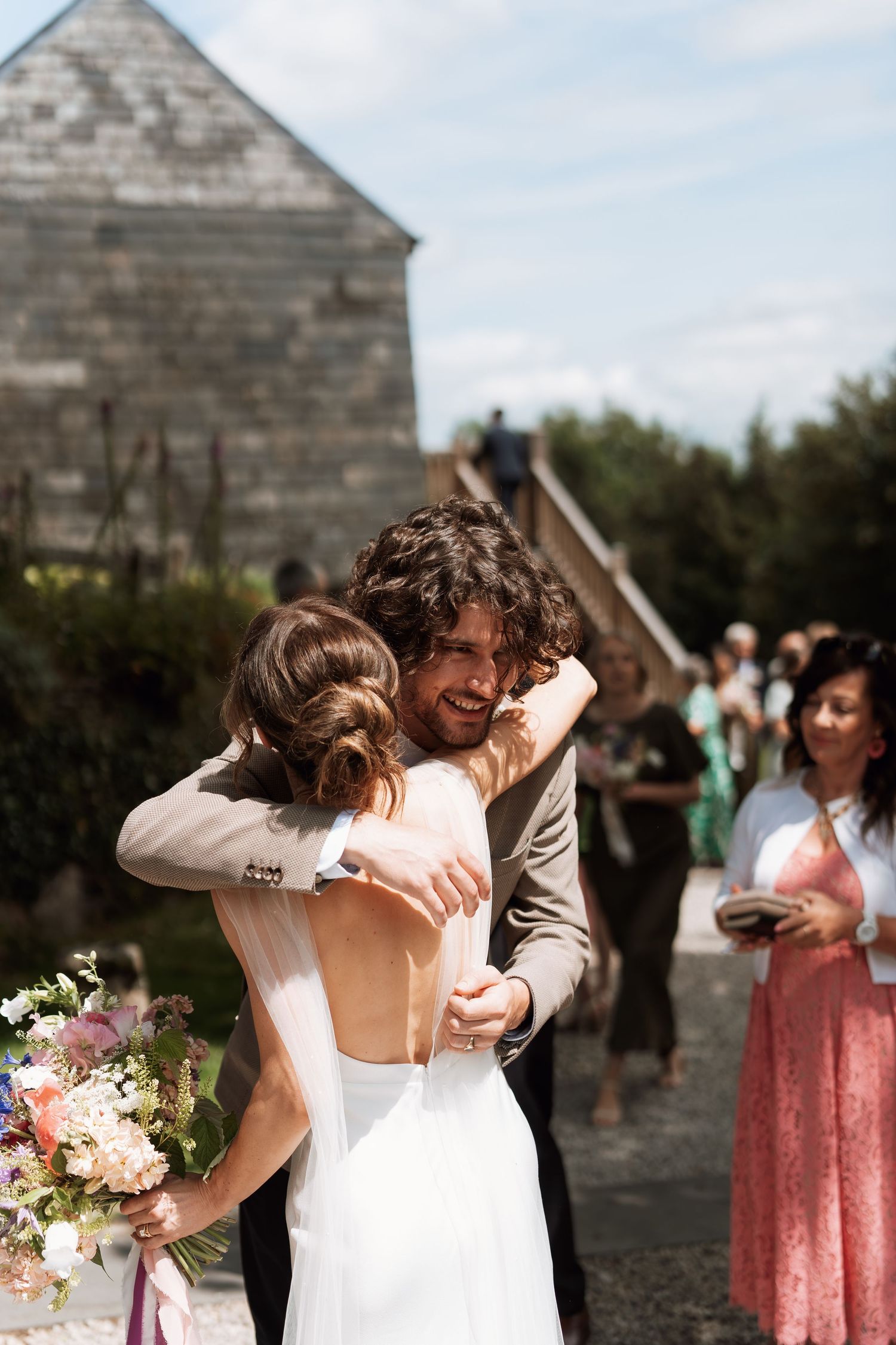 Wedding couple sharing emotional embrace outside stone building with guests watching.