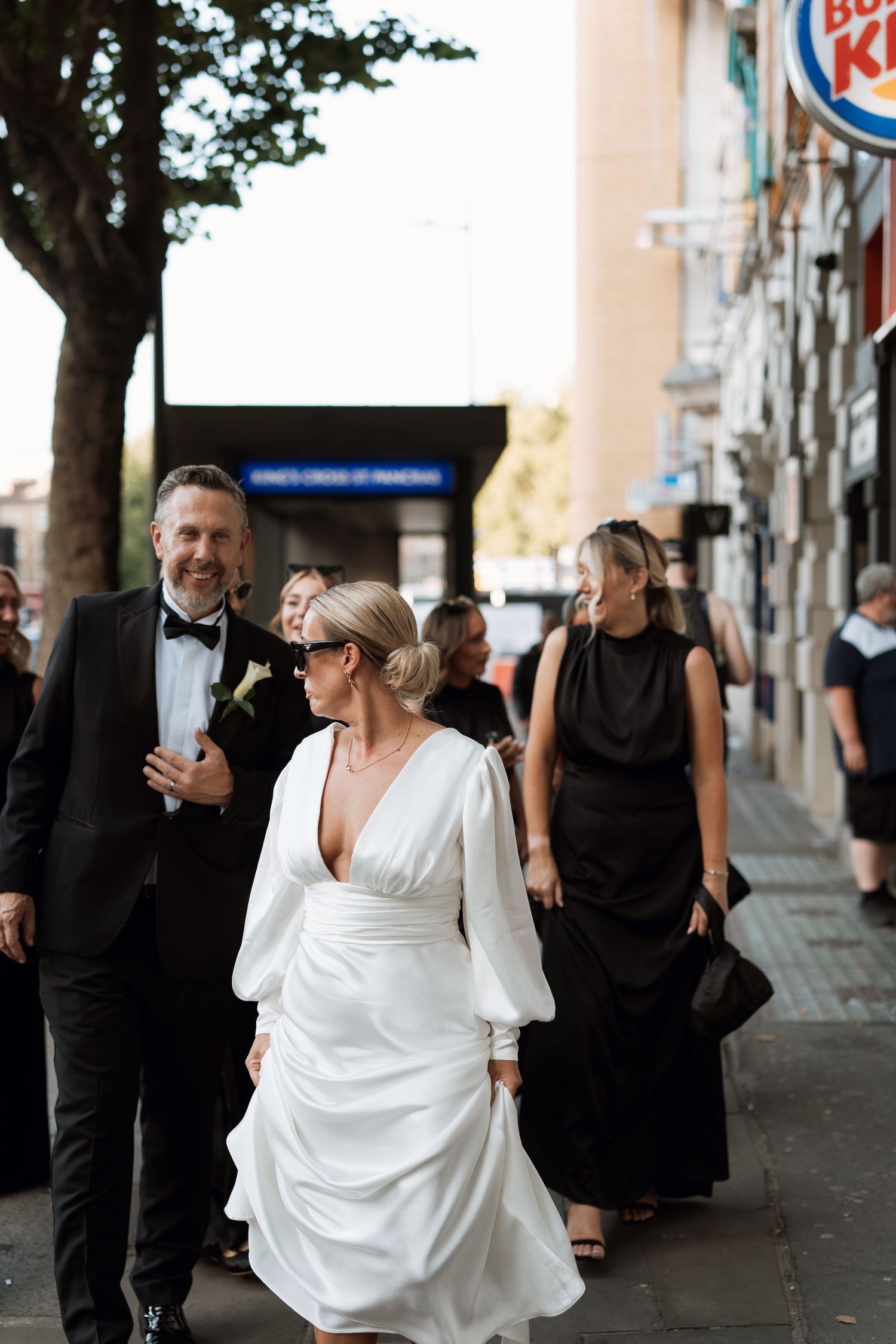 Wedding party walking down city sidewalk with bride in white dress and guests in black attire.