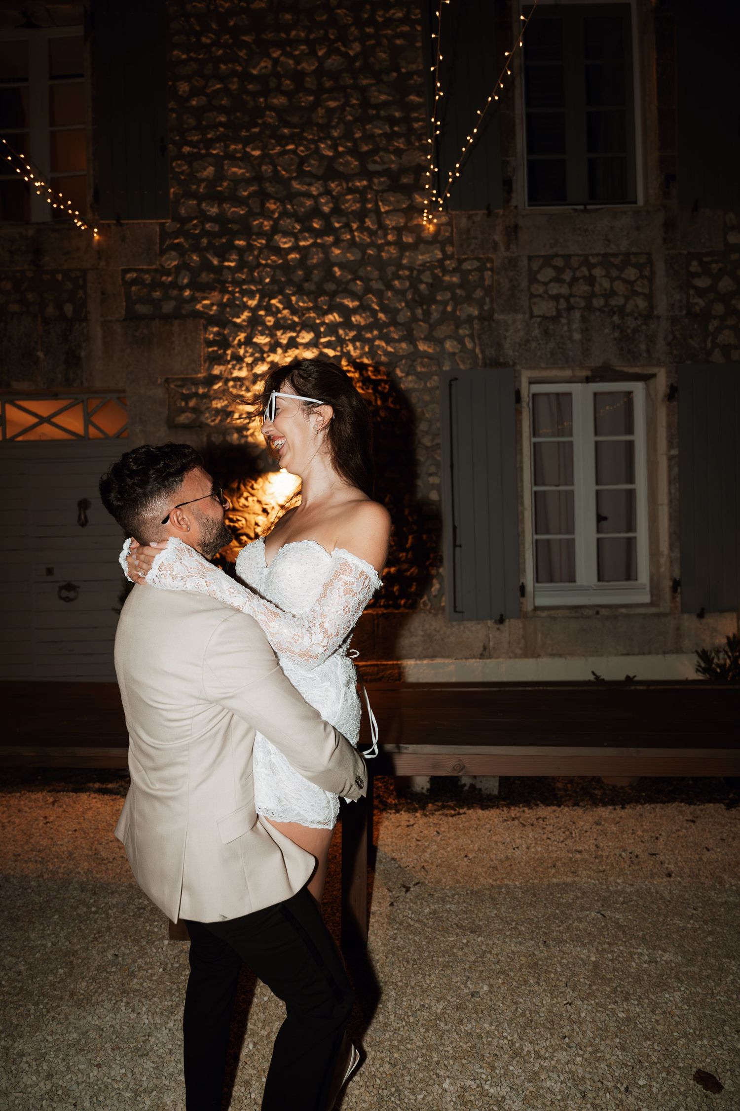 Romantic nighttime dance between newlyweds under warm string lights against stone building backdrop.