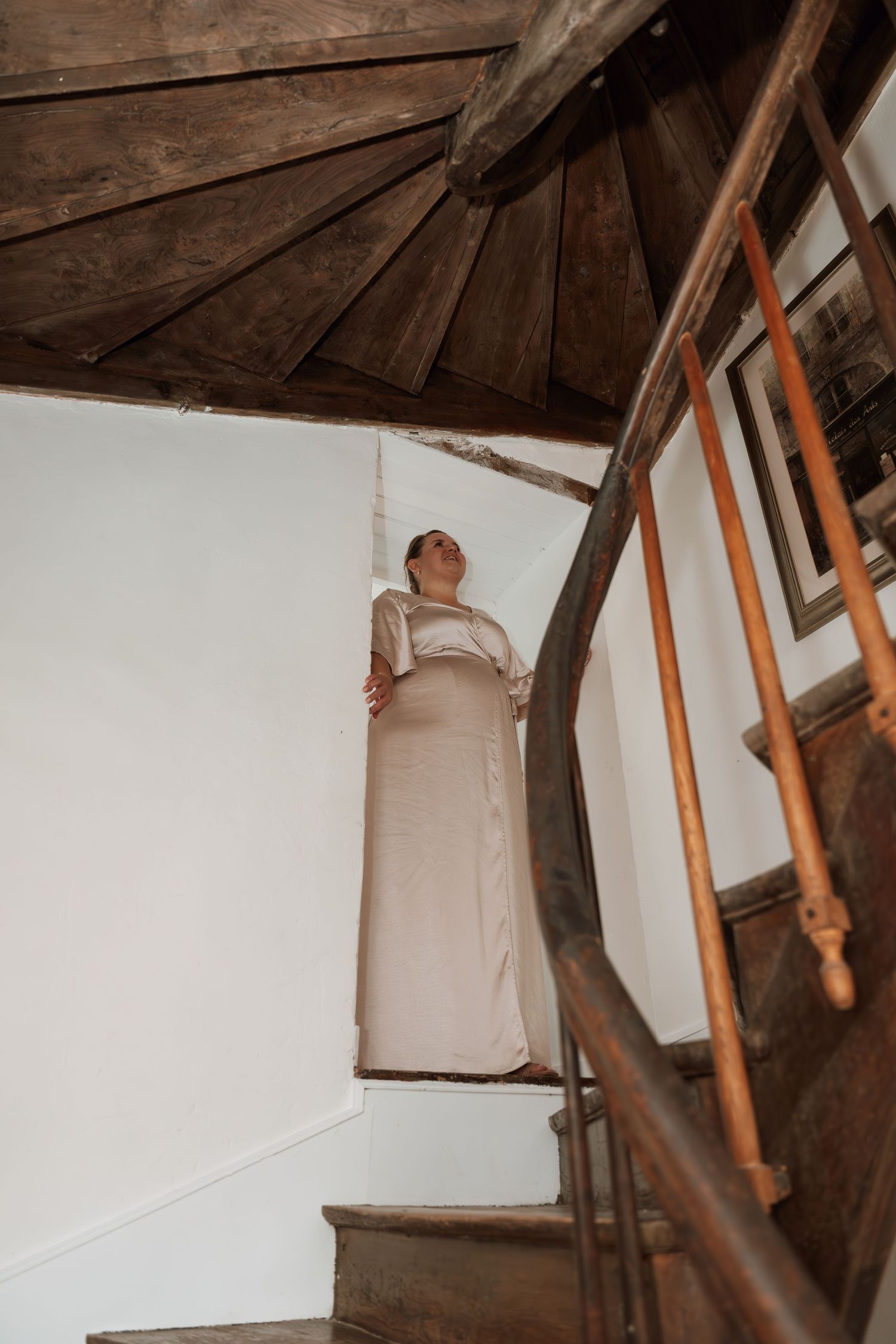A figure in a cream-colored dress stands on wooden spiral staircase in an old building.