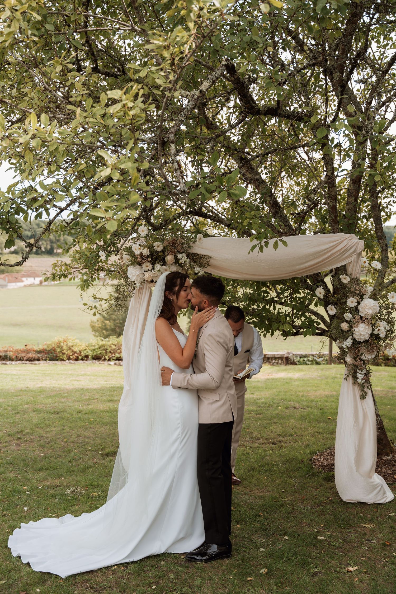 A romantic outdoor wedding ceremony takes place under trees with white fabric draping.