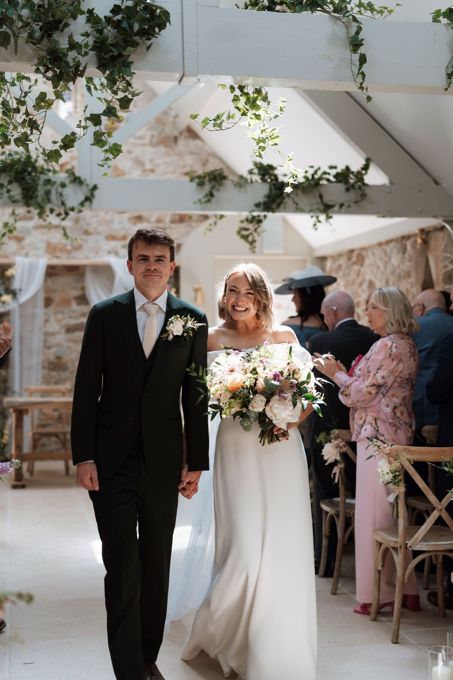 Bride and groom walking down the aisle in a rustic barn venue adorned with ivy, bride holding a pastel bouquet.