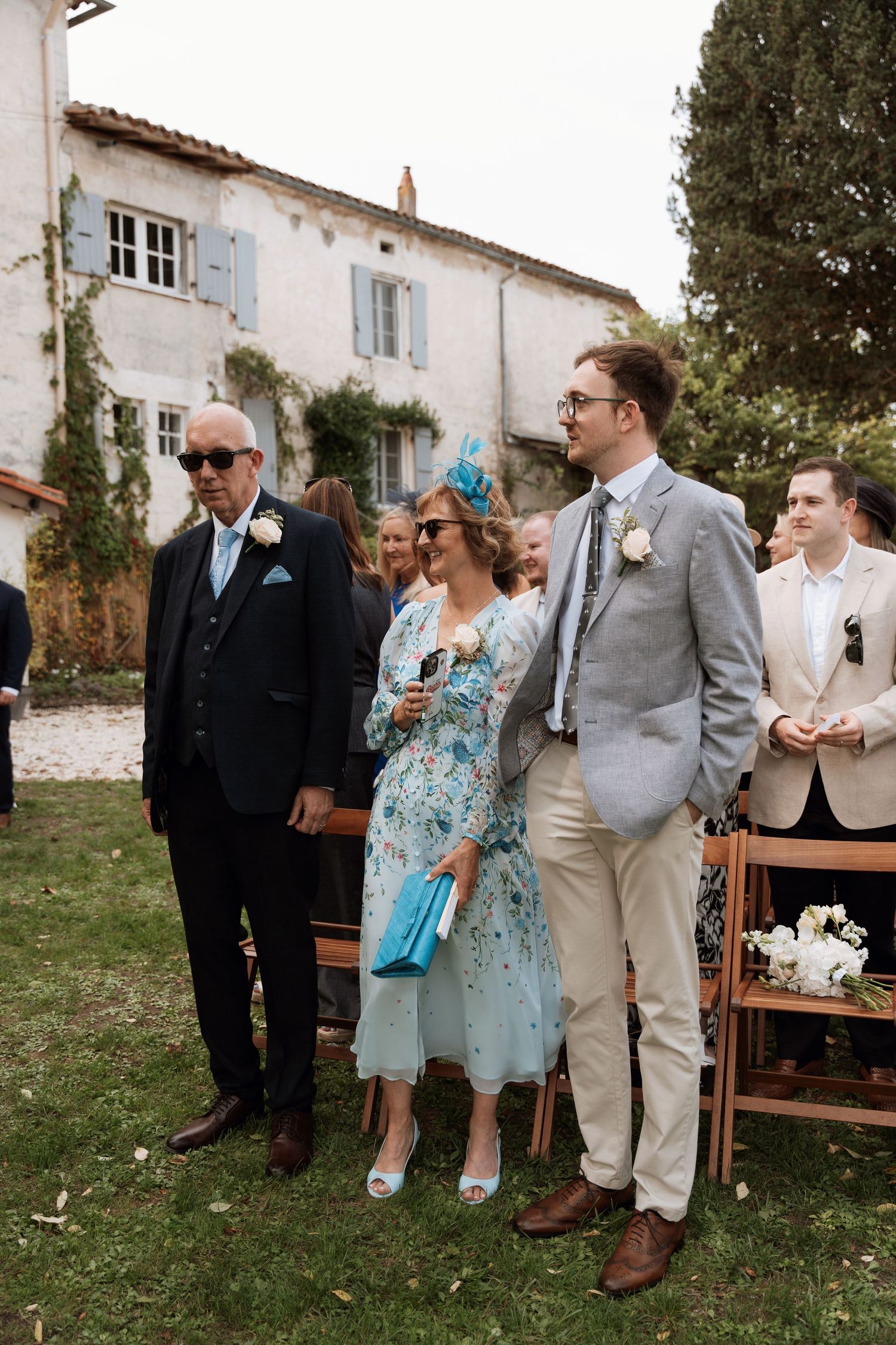 Wedding guests in formal attire gathered outside a rustic stone building.