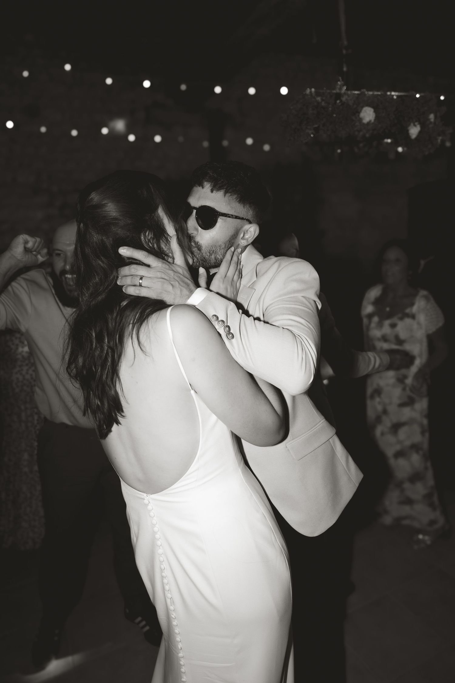 Black and white photo of couples dancing under string lights at an evening wedding reception.