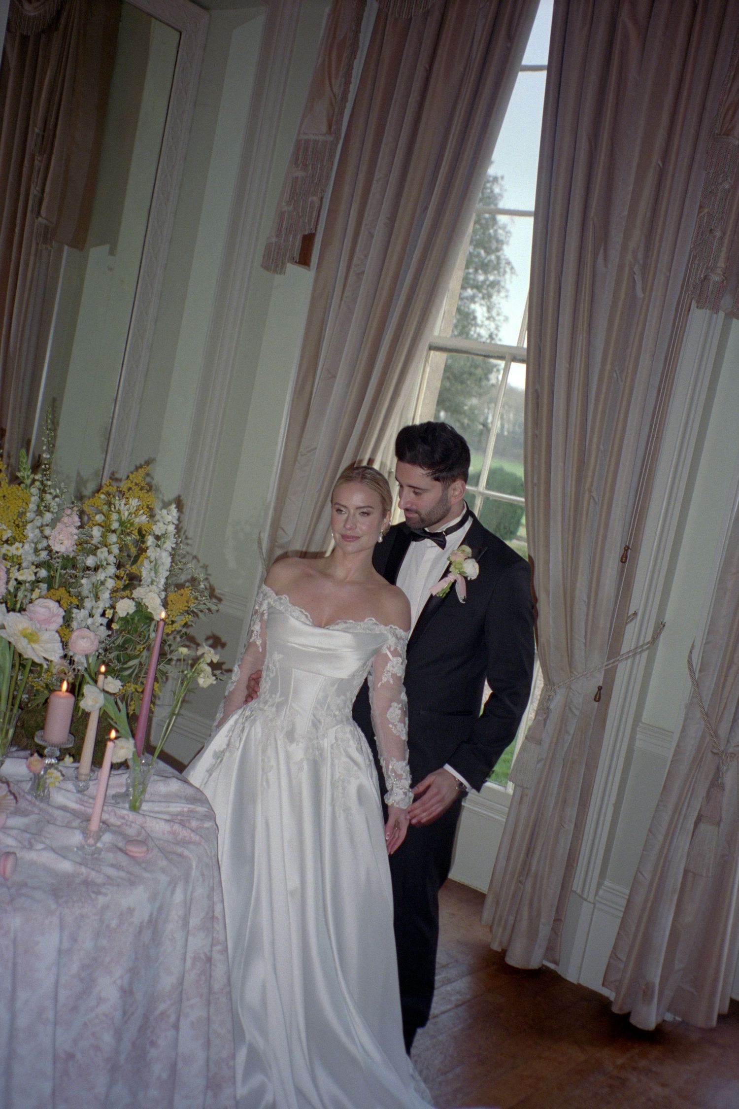 A couple in formal wedding attire stand together in an elegant room with tall windows and flowing pink curtains.
