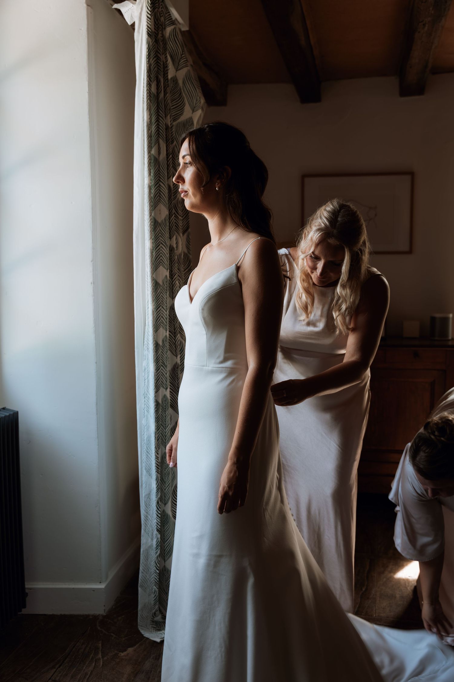 Women in white dresses stand near a window in dramatic lighting.