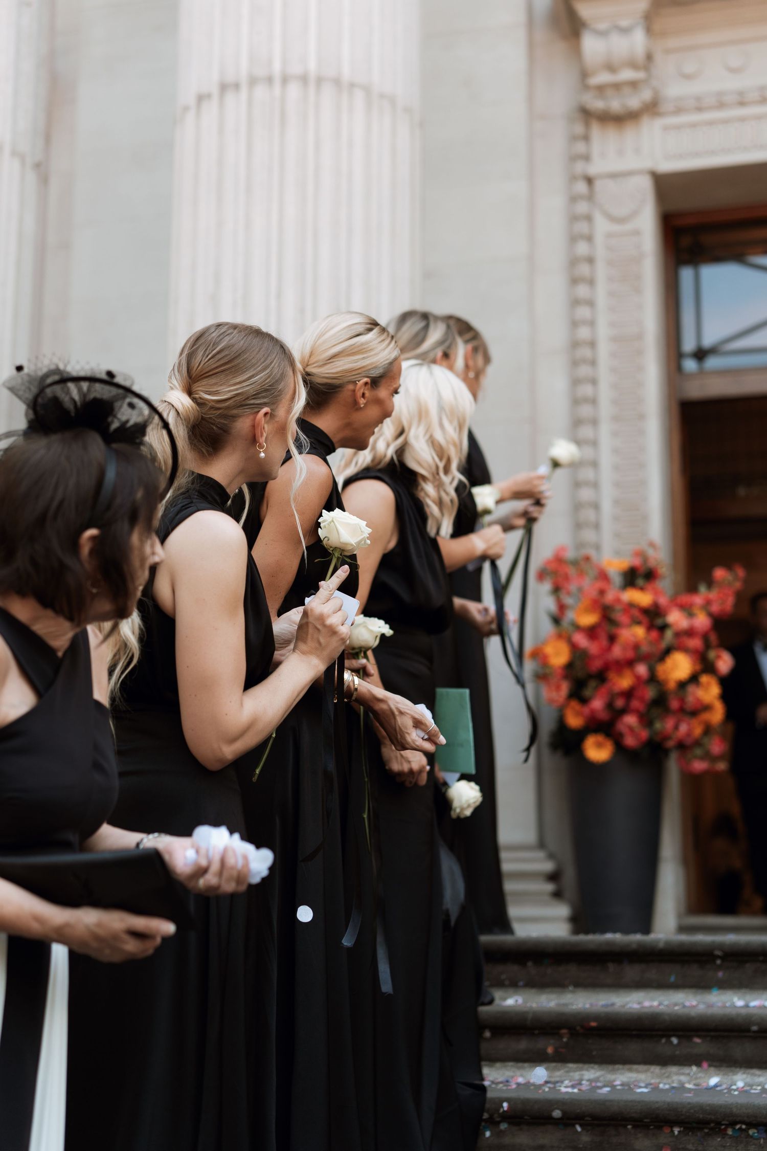 Bridesmaids in black dresses line up holding white bouquets on elegant stone steps.