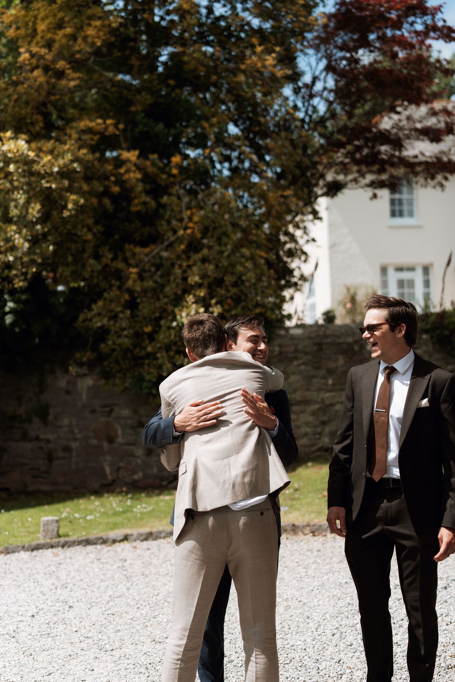 Wedding party members in formal attire embrace and celebrate on a gravel path outside.