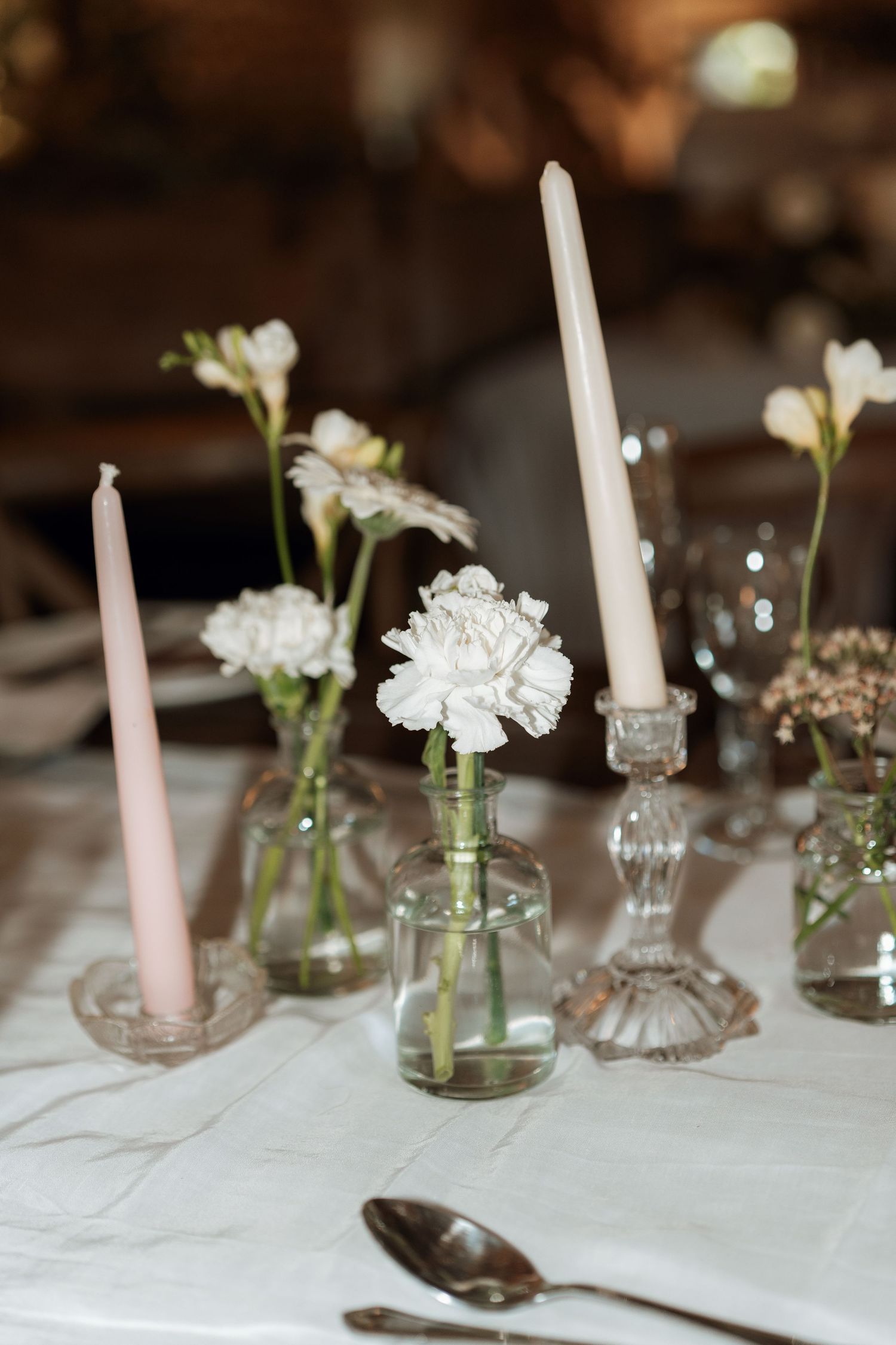 Elegant wedding table decor with white flowers in small glass vases and tall white candles on a white tablecloth.