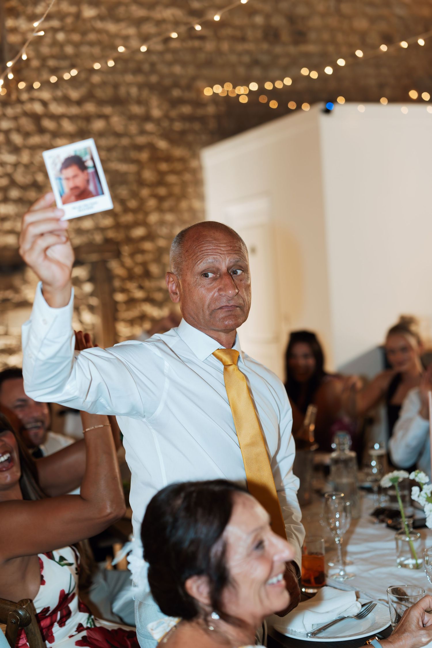 Wedding guest in light colored shirt and yellow tie holds up photos during reception celebration.